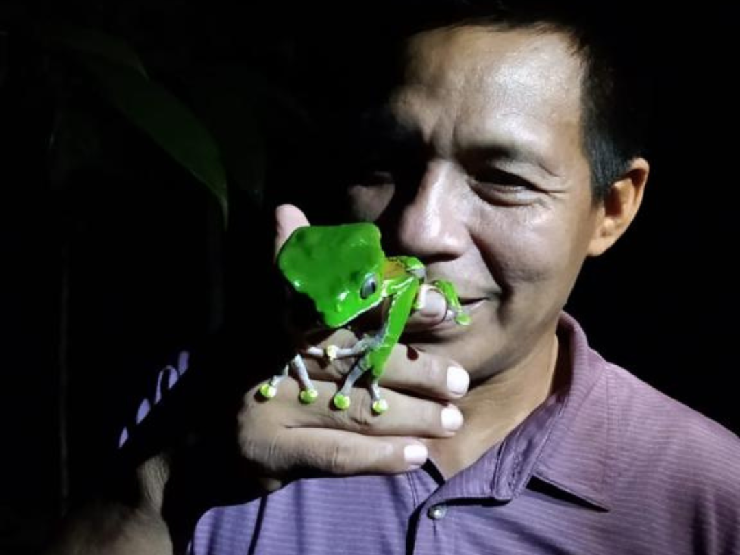 A man with short dark hair, wearing a purple collared shirt, holding a Kambo Tree Frog close to his face. The background is dark.