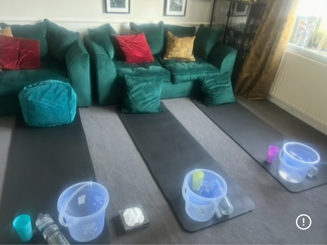 Three black mats with plastic buckets, cups, and tissues, set up on a carpet in front of a green sofa with colorful pillows in a living room.