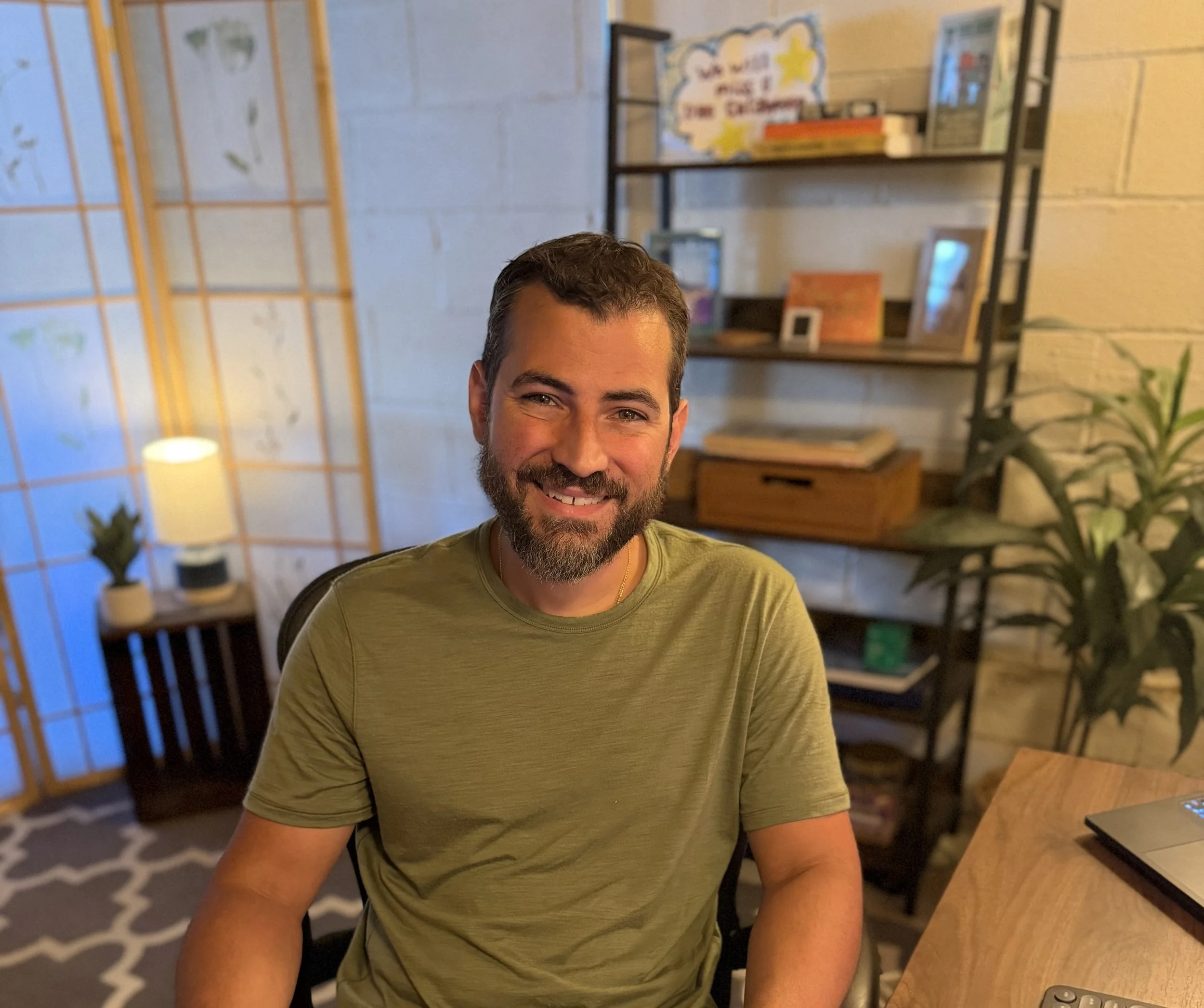 A smiling man with dark hair and a beard sitting in a cozy room with a bookshelf, plants, and warm lighting.