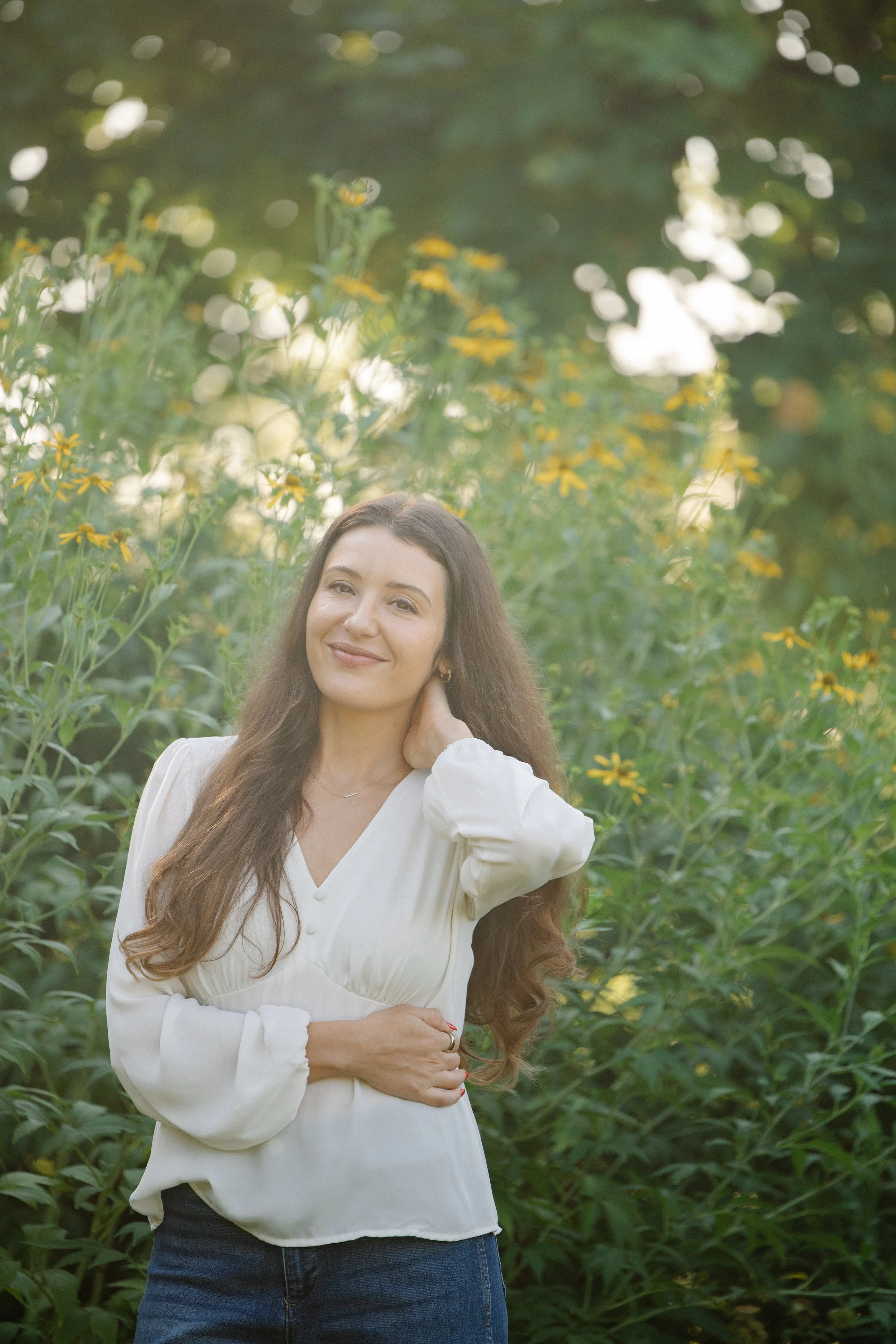 A woman with long brown hair wearing a white blouse and dark jeans is standing outdoors in a lush garden with yellow and white flowers, smiling and touching her neck.