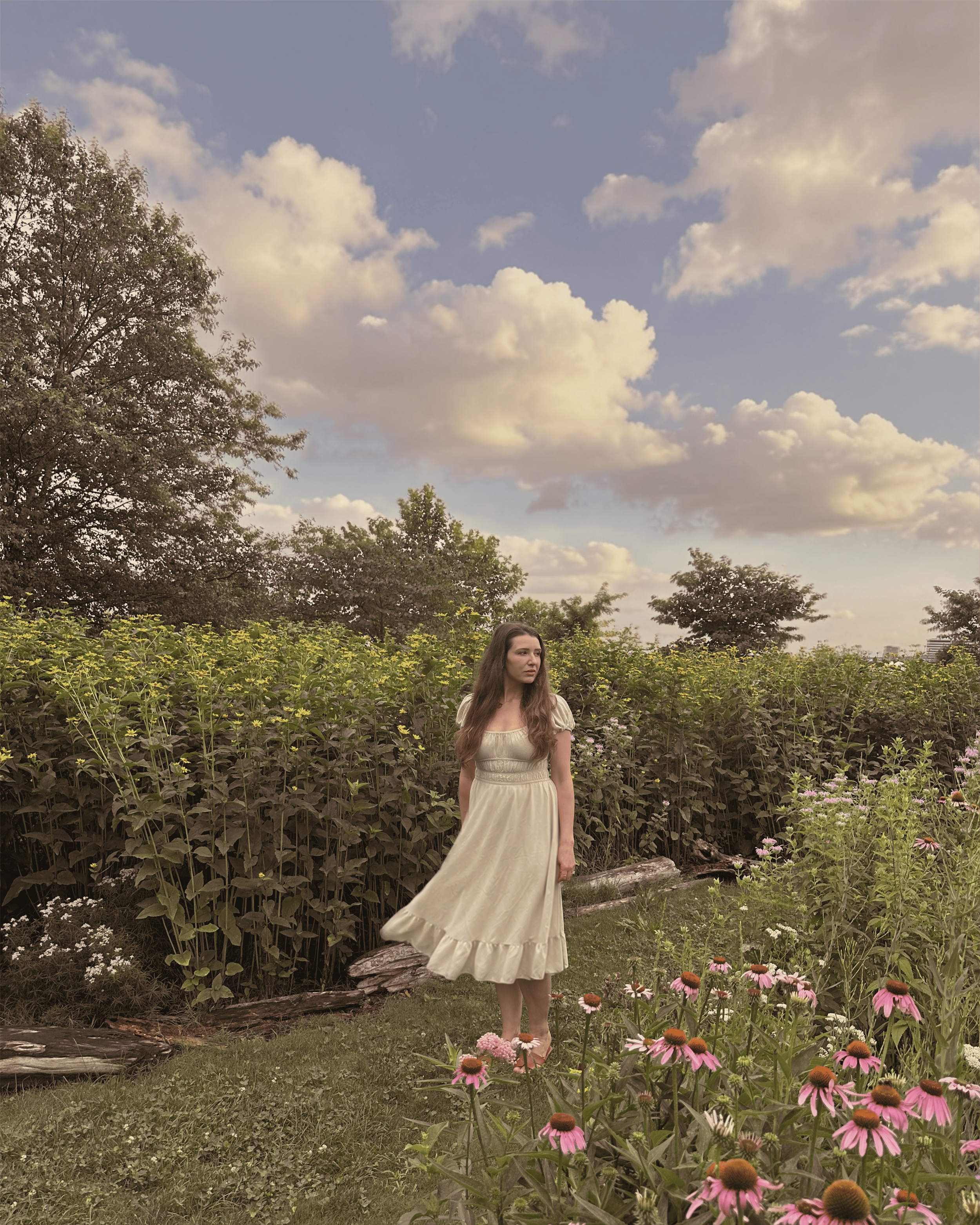 A woman with long, wavy brown hair wearing a cream-colored dress standing in a garden filled with pink and purple flowers. Behind her is lush green foliage and trees, with a blue sky and scattered clouds above.