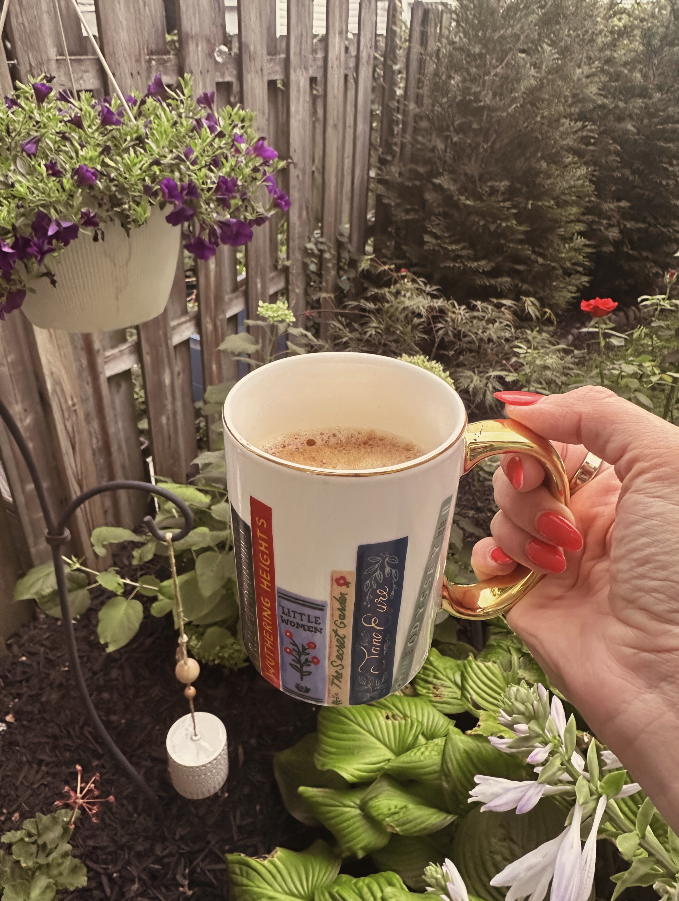 A close-up of a person holding a white ceramic mug with a gold handle, filled with a hot beverage, in a garden setting with purple and white flowers, green foliage, a wooden fence, and garden ornaments in the background.