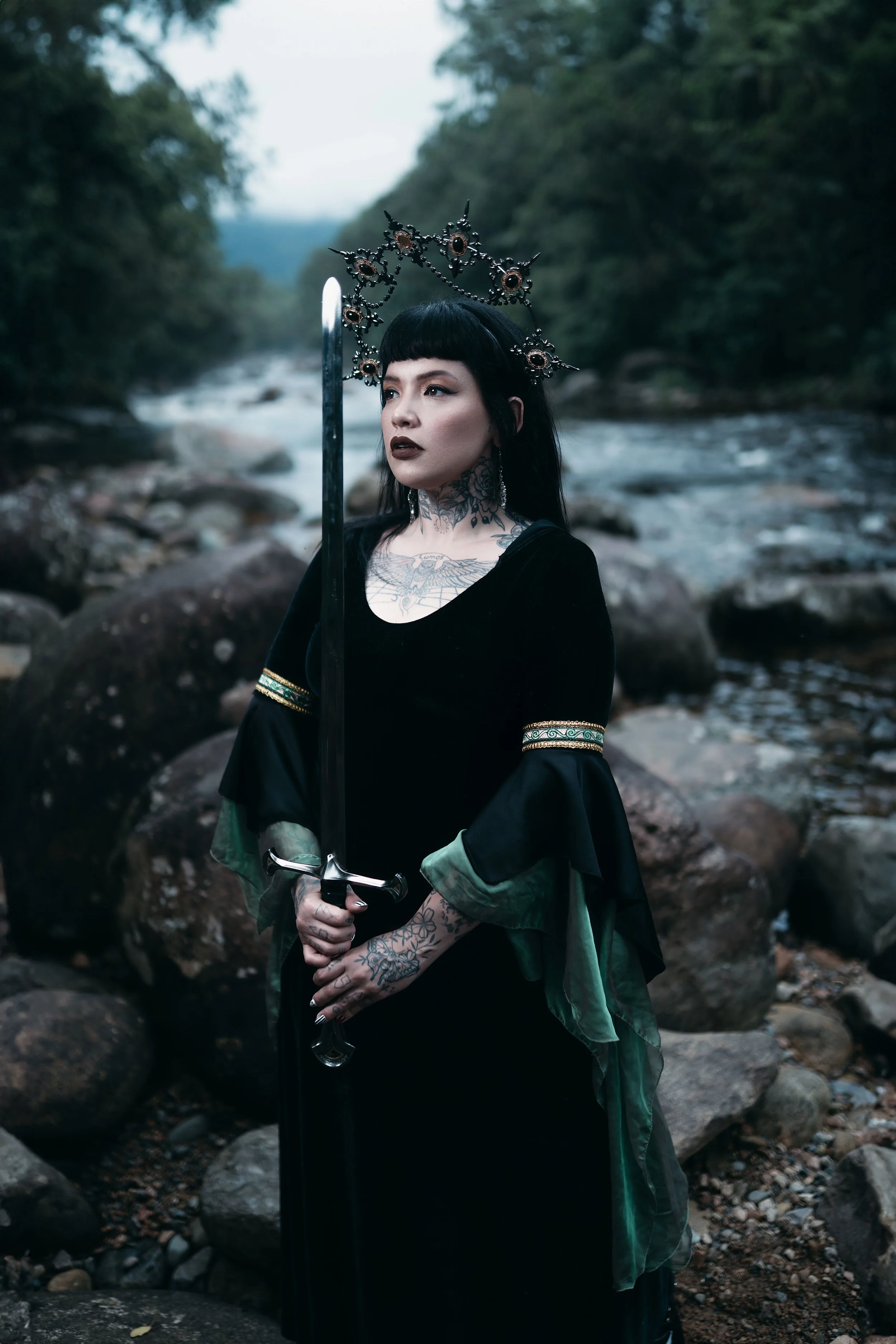 A woman dressed in black with tattoos and gothic makeup, wearing a black and green dress and ornate crown, stands in a rocky stream holding a sword, with a forested background.