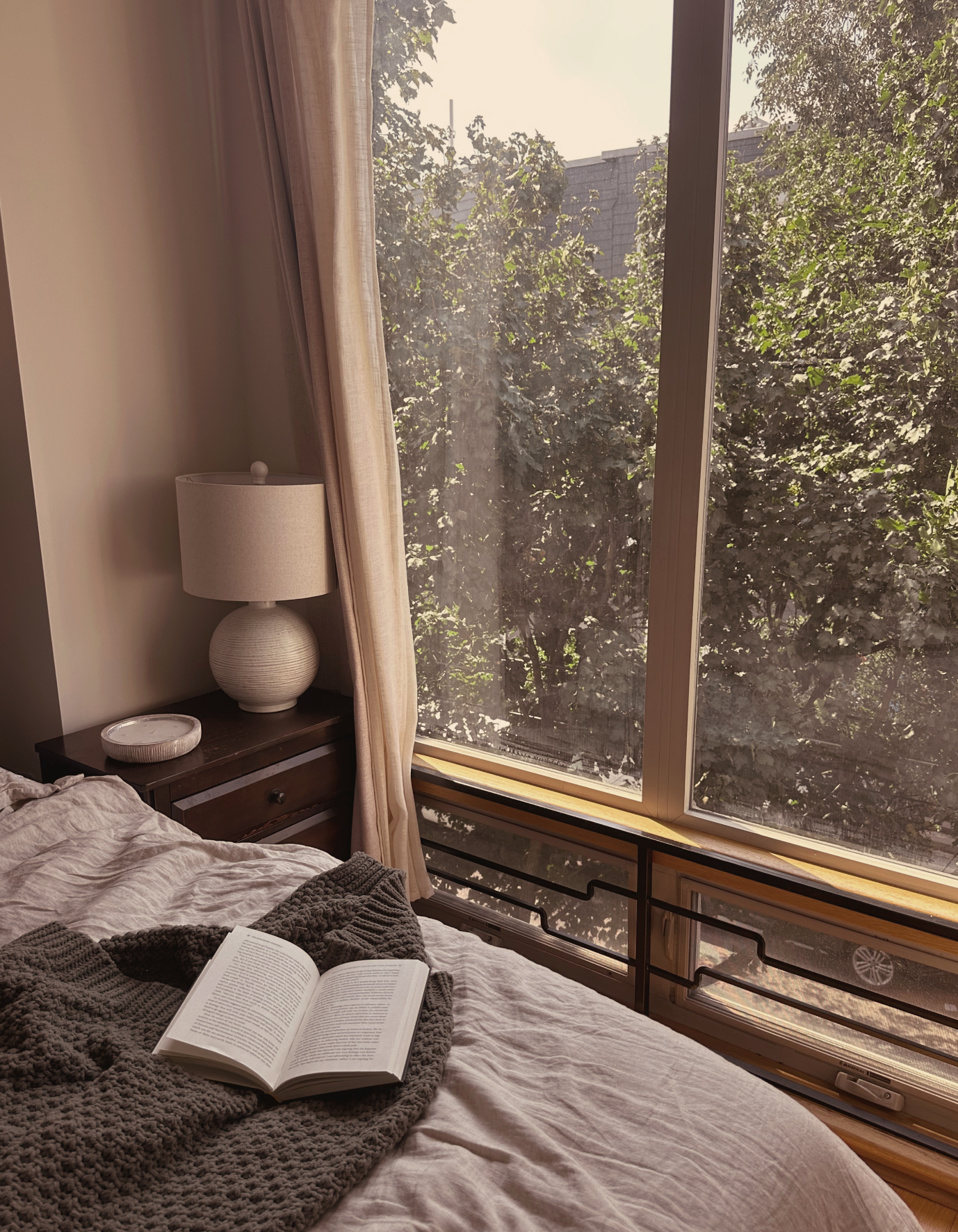 A bedroom with a large window showing trees outside, a bedside table with a white lampshade, an open book on the bed, and a cozy blanket.