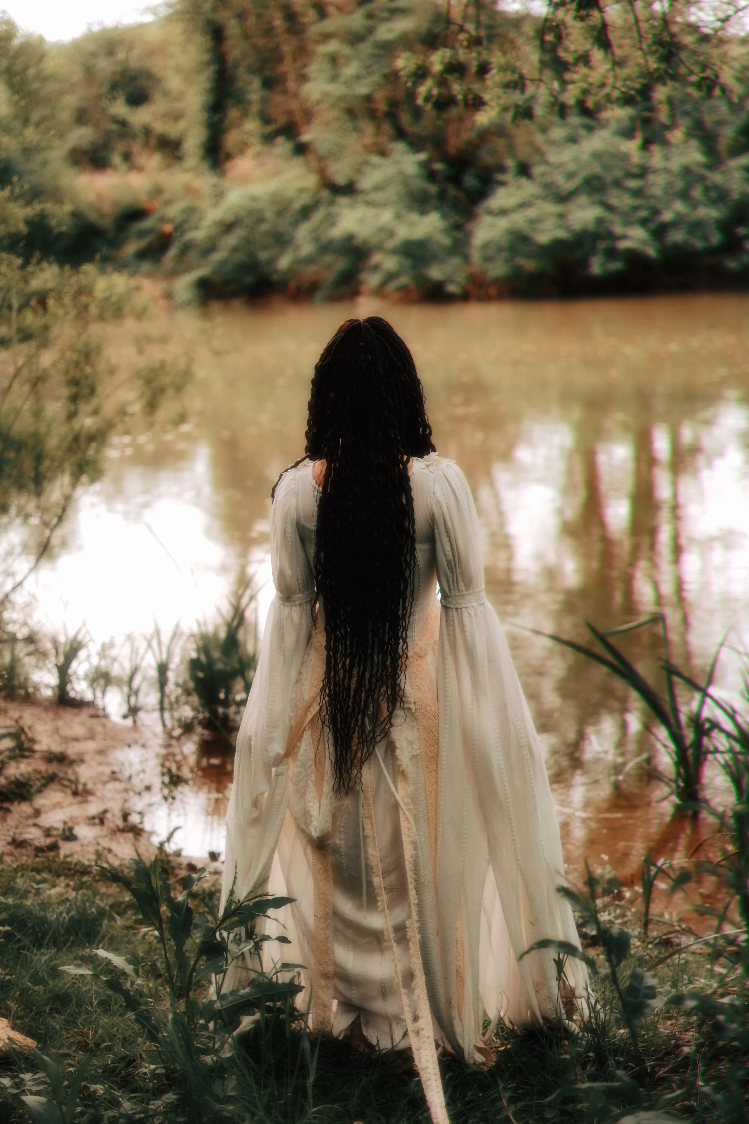 A woman in a vintage white dress with long, dark, curly hair stands by a river in a lush, green forest, facing away from the camera.
