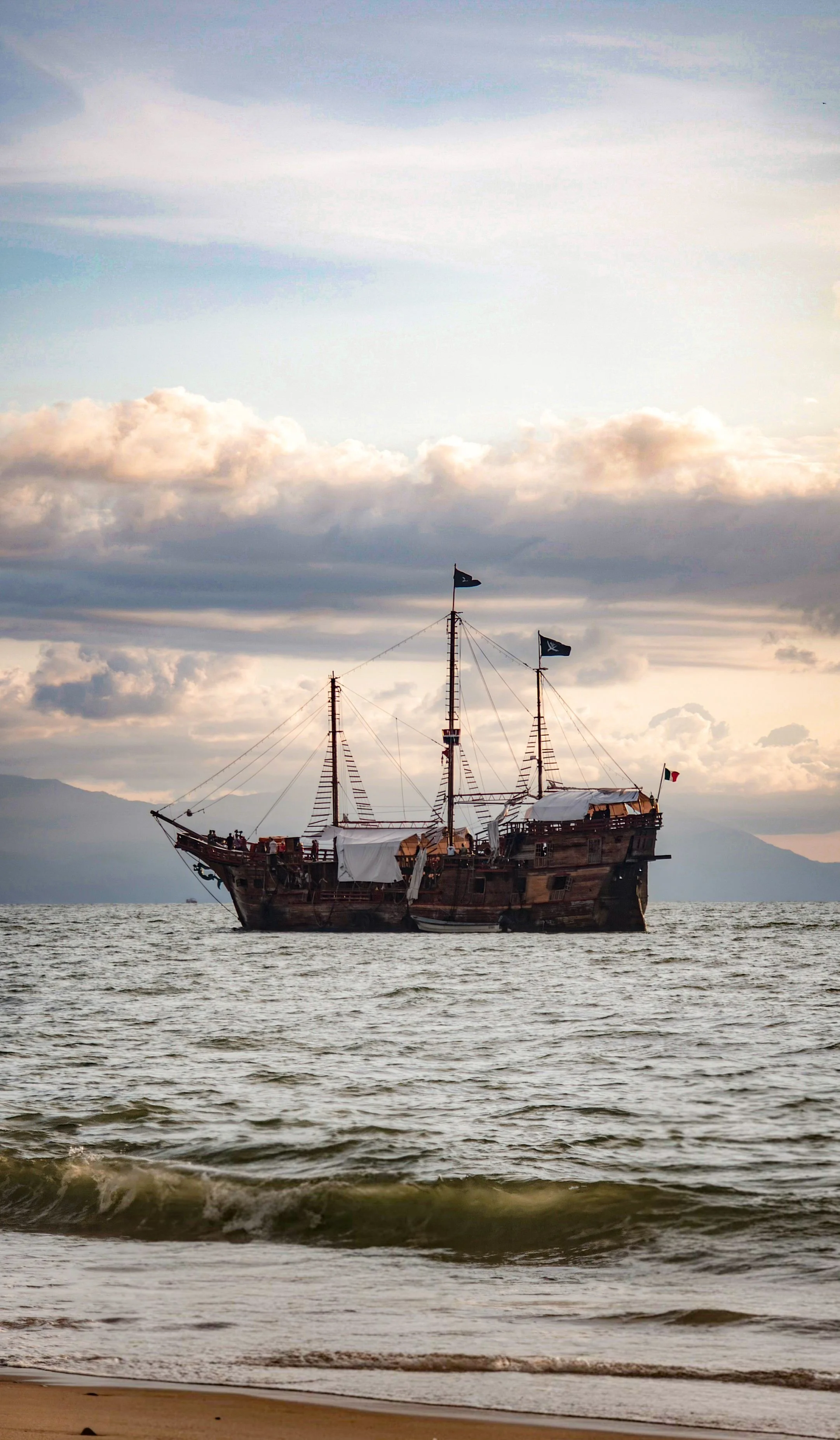 A large, wooden pirate ship with black flags decked with skull and crossbones, sailing on the ocean with a cloudy sky and distant mountains in the background.