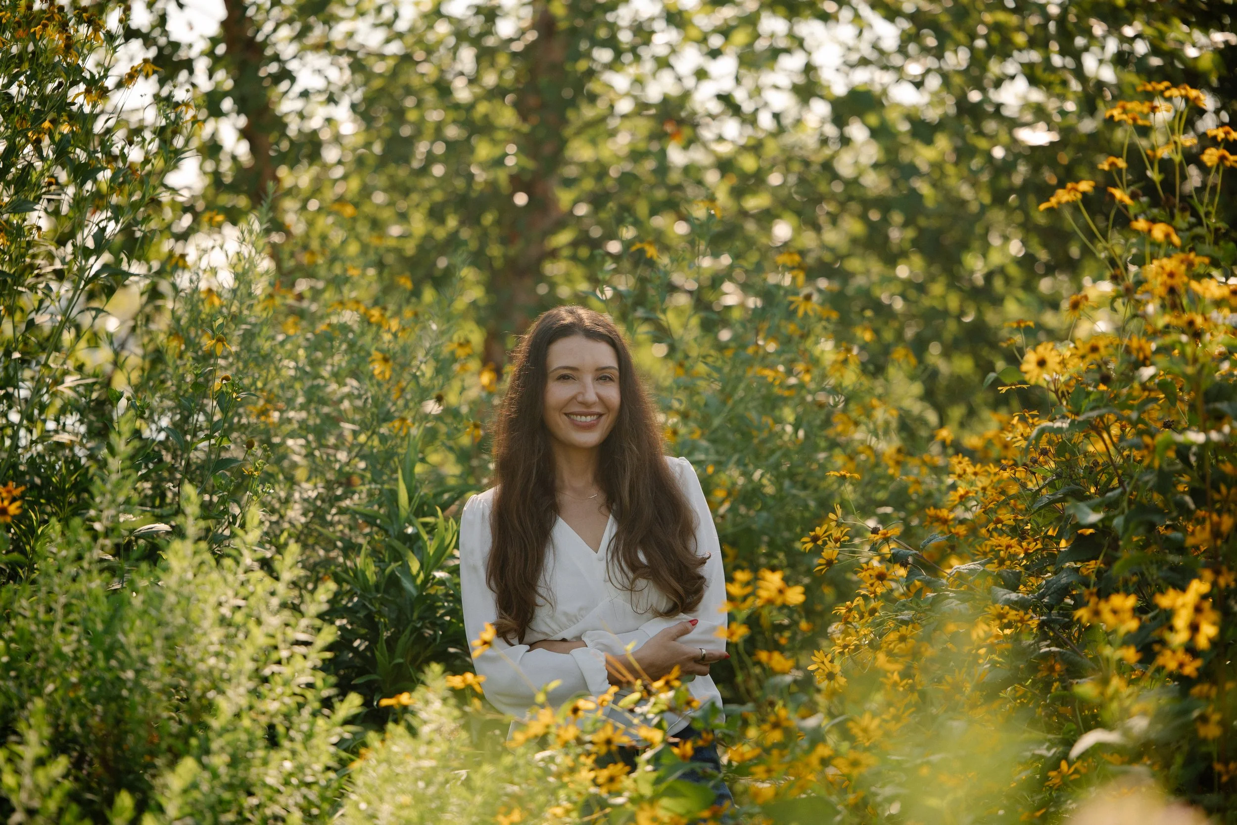 A woman with long brown hair smiling and standing with arms crossed amidst yellow flowers and greenery on a sunny day.