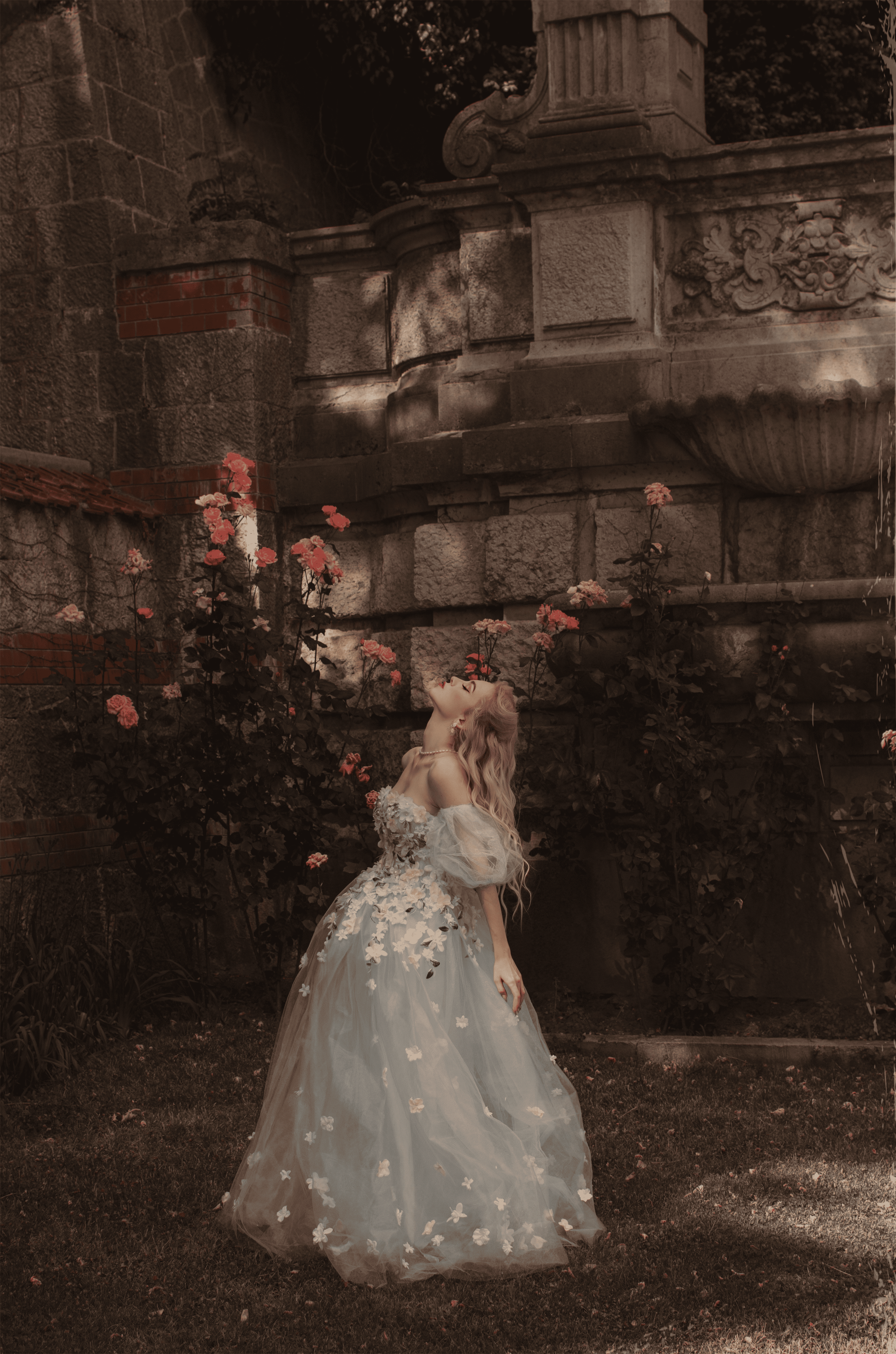 A woman with long, wavy blonde hair wearing an off-shoulder, pastel gown with floral appliqué stands outdoors near a stone wall with roses and greenery, and a fountain in the background. She is looking up with a serene expression, surrounded by a lush garden setting.