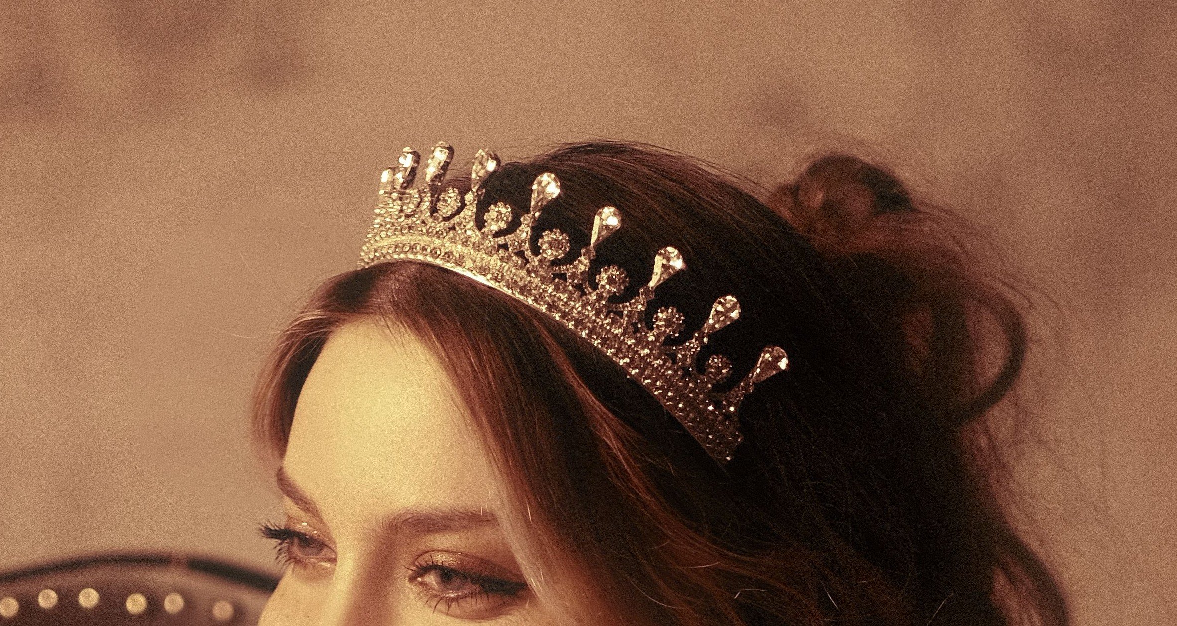 Close-up of a woman's face with a tiara on her head, with brown hair styled in waves.