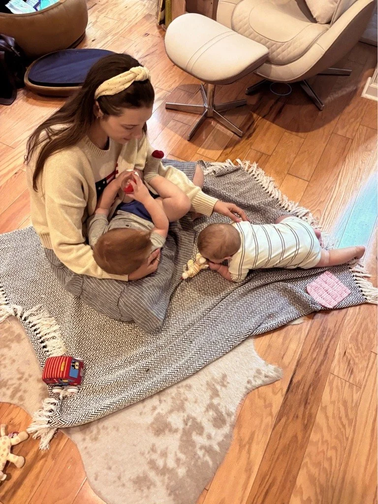 A woman sitting on a blanket on a hardwood floor with three young children, two lying down and one crawling. Toys are scattered around, and there is a beige chair and a pet bed in the background.