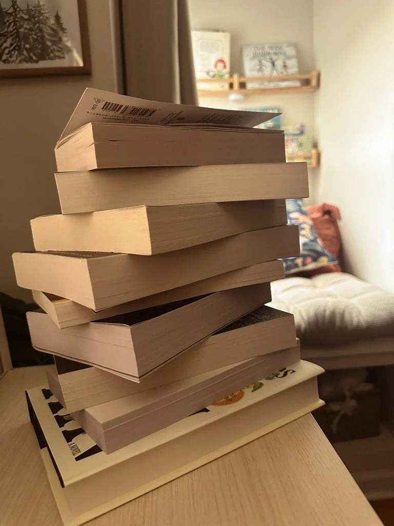 A stack of hardcover and paperback books on a wooden surface in a cozy room with a beige wall, a sofa with pillows, and floating shelves with books in the background.