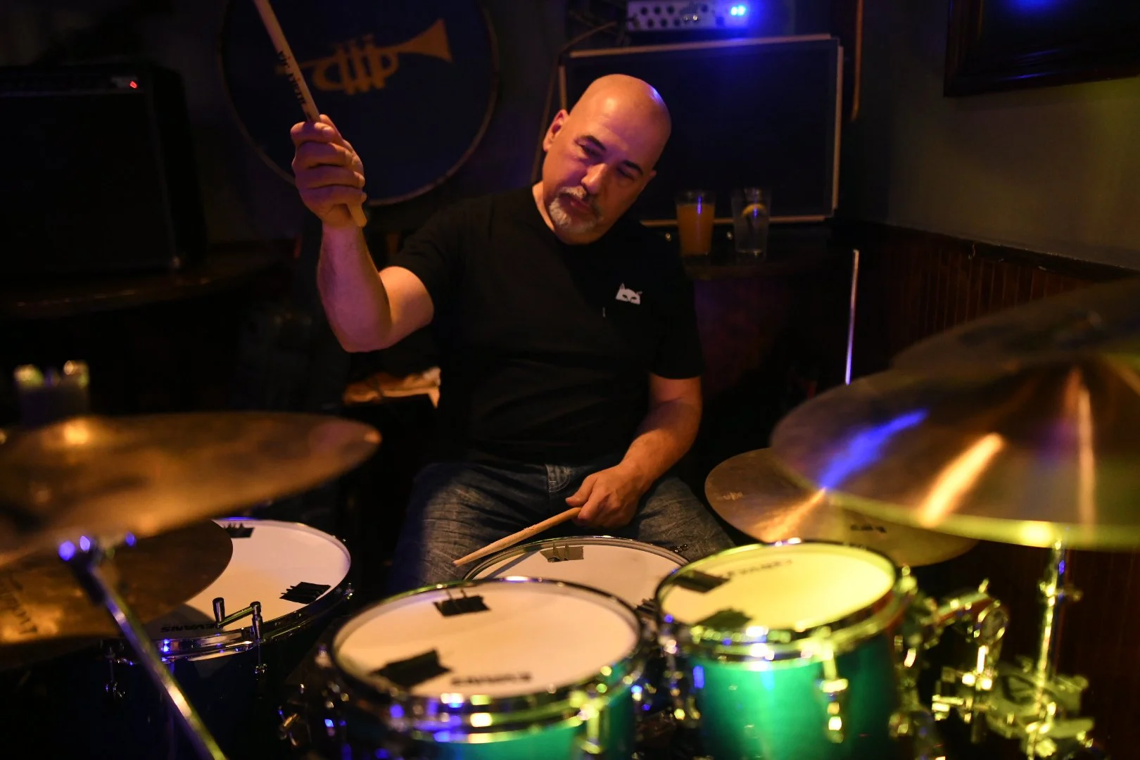 A man playing drums in a dimly lit room, holding drumsticks, with various drums and cymbals around him.