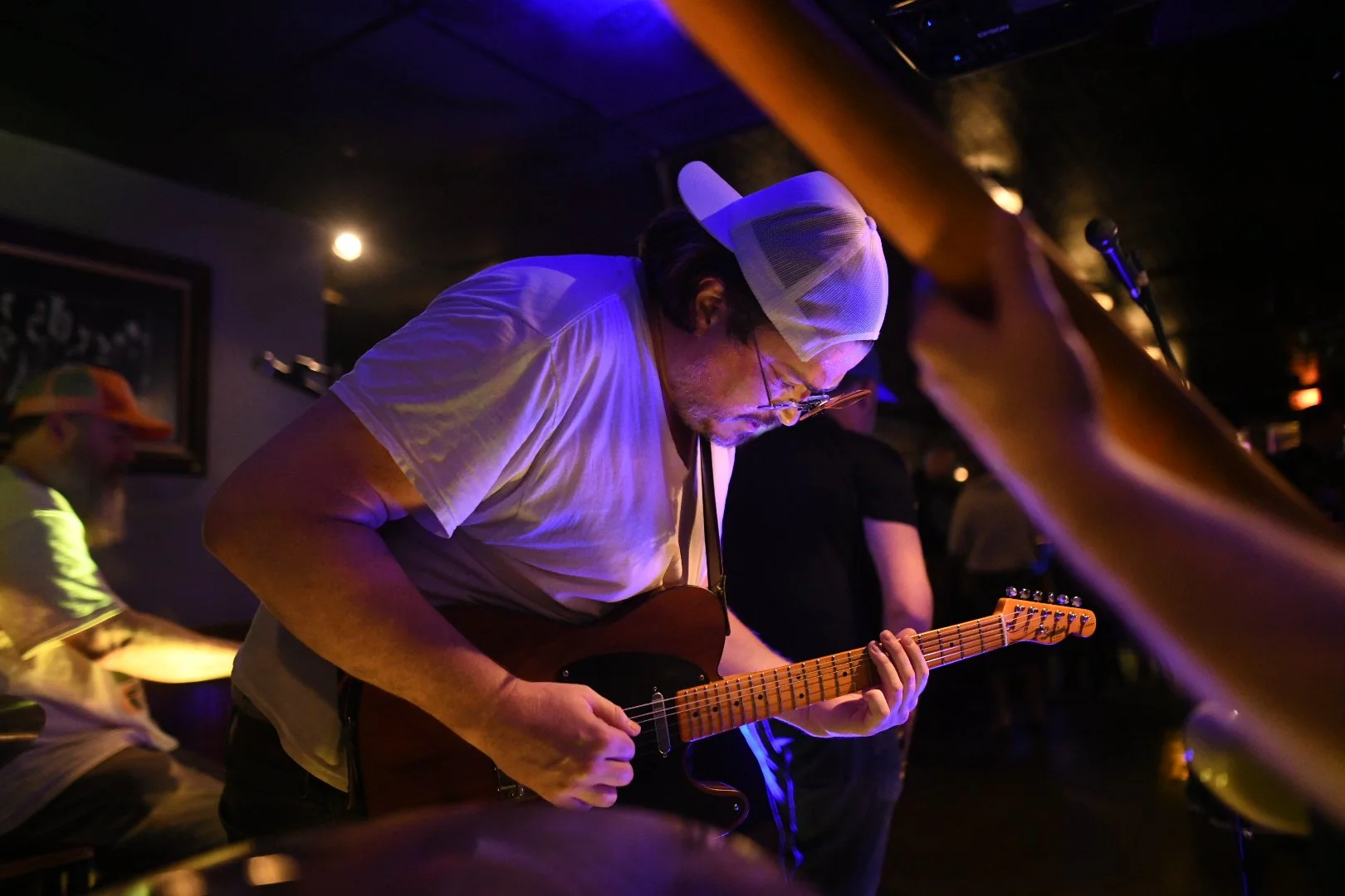 A man wearing a white cap, glasses, and a white t-shirt playing an electric guitar in a dimly lit room with a dark ceiling and some visible patrons in the background.
