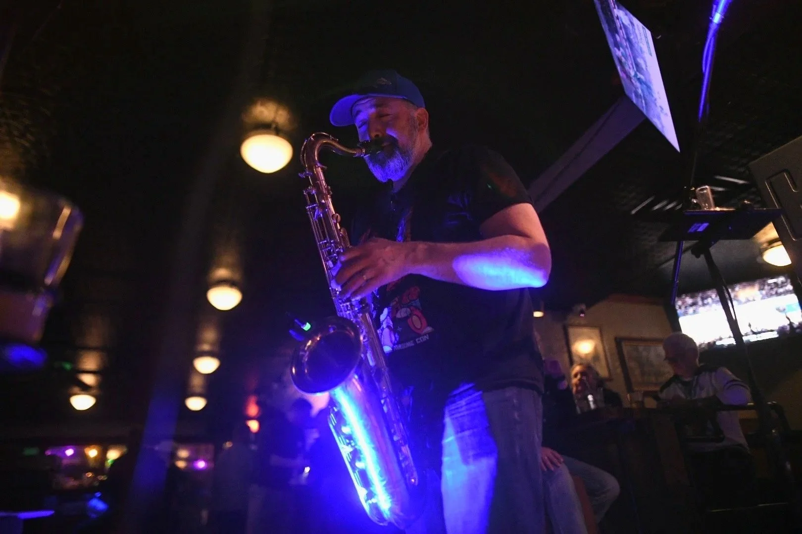 A man playing a saxophone with purple and blue lighting in a dimly lit bar or club. There are patrons sitting in the background watching.