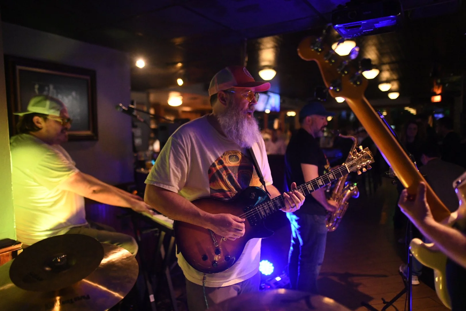 Three musicians performing live in a dimly lit bar. The central figure is a bearded man wearing a red cap, orange-tinted glasses, and a T-shirt with a colorful graphic, playing an electric guitar. To his left, a person wearing a beige cap and shirt is playing the keyboard, and to his right, a saxophonist in a dark cap and shirt is playing saxophone. The background features warm lighting, a framed picture on the wall, and a crowd of listeners.