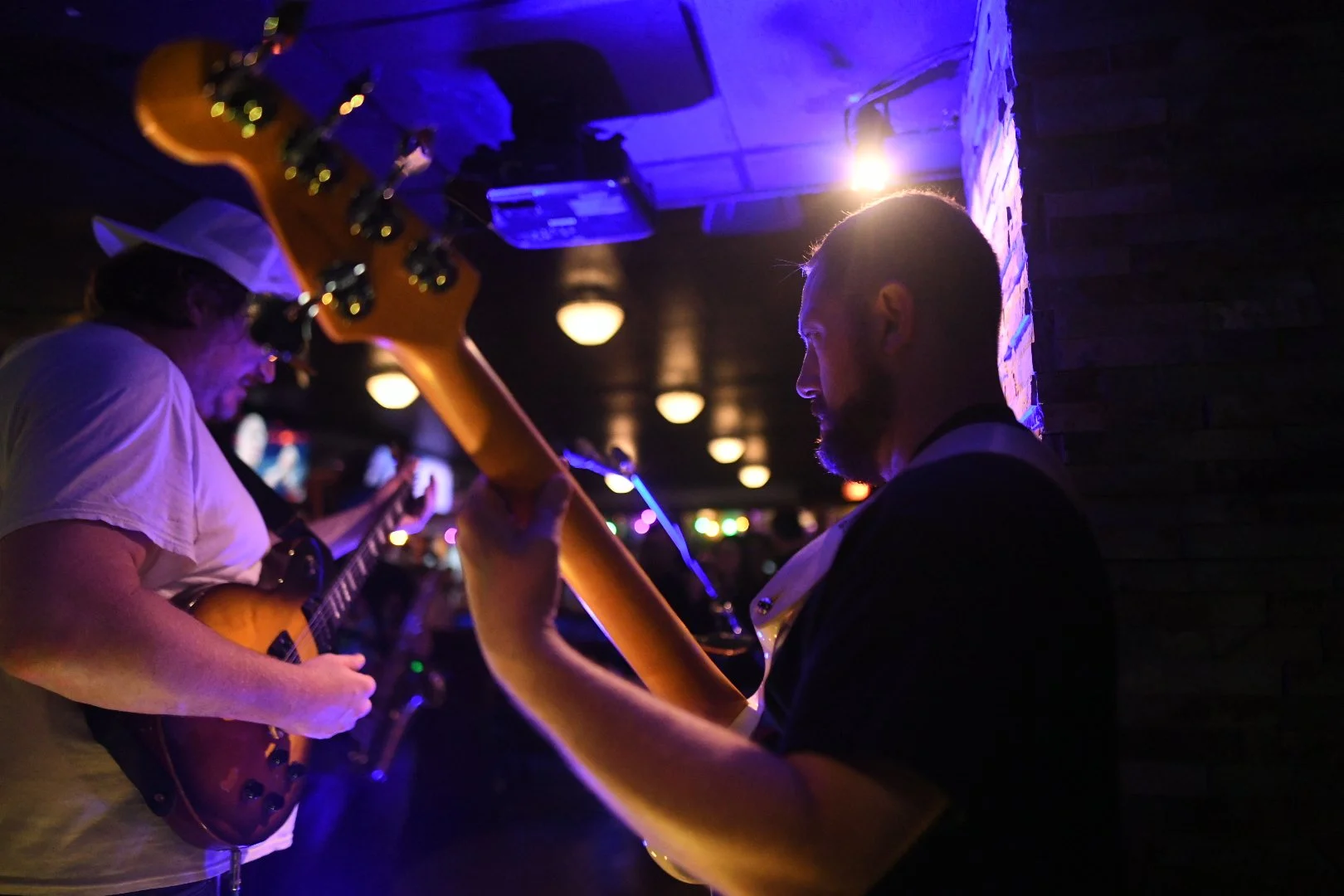 Two musicians playing guitars in a dimly-lit venue with purple and yellow lighting. One is wearing a white shirt and a white hat, the other has a patterned shirt and short hair. There are blurred colorful lights in the background.
