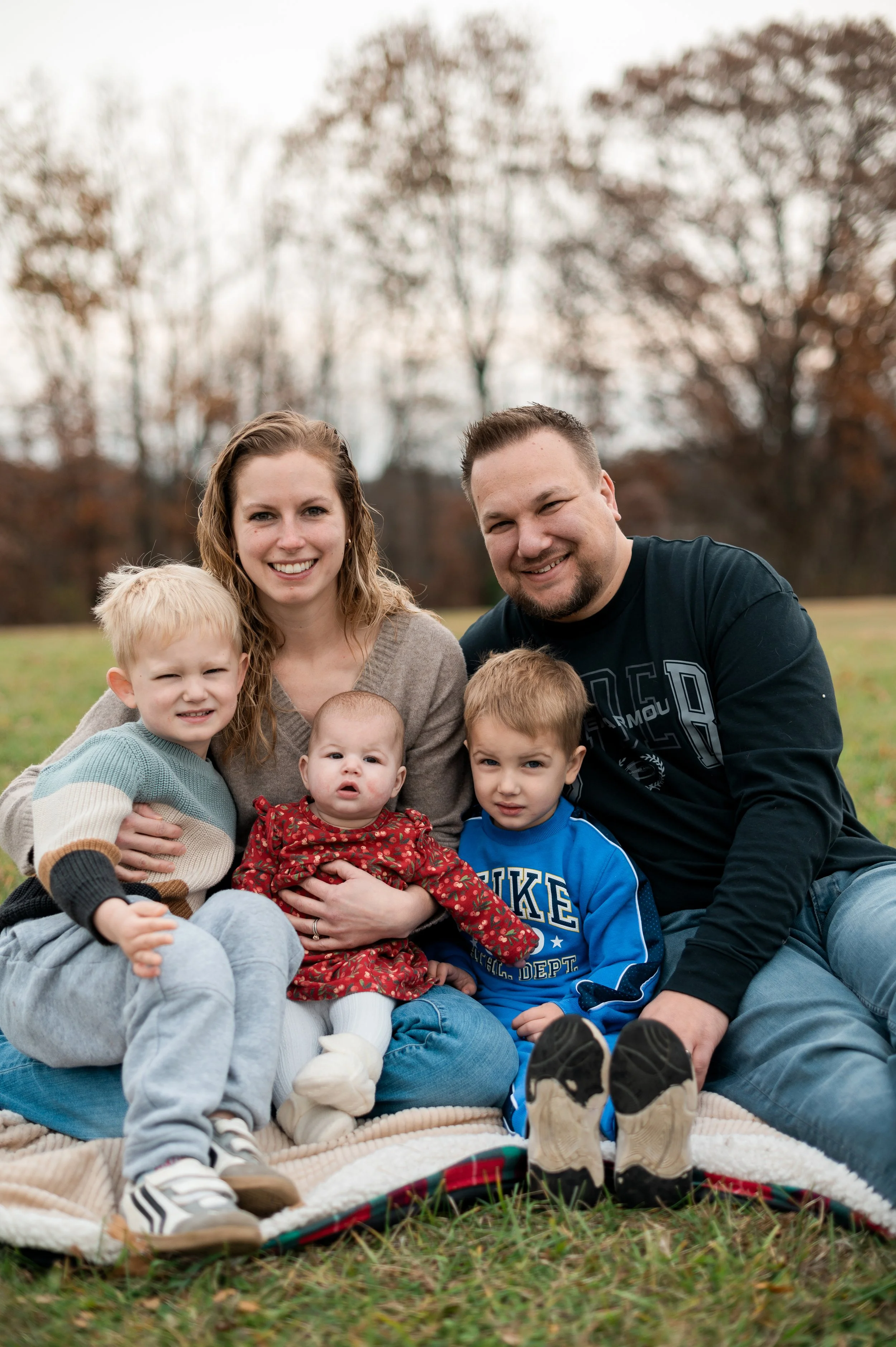 A family of six sitting on a blanket in a park with autumn trees in the background. The group includes a woman, a man, and four children, and they are smiling and looking at the camera.