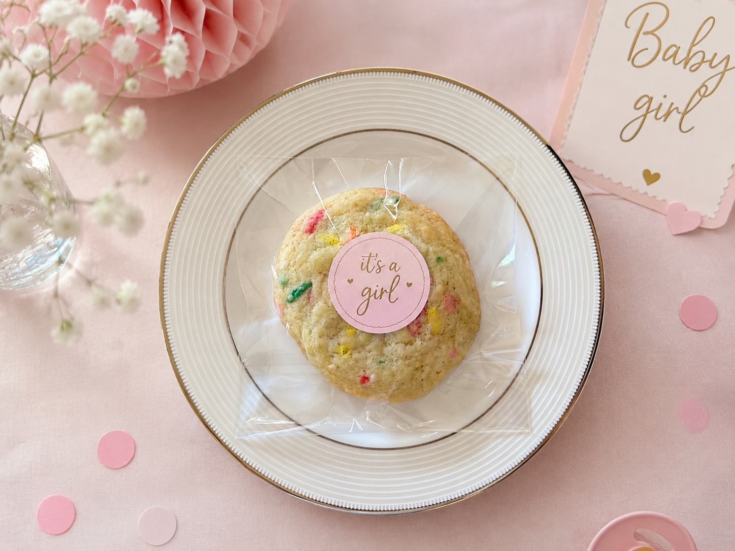 A table with a pink-themed baby girl celebration setup including a flower bouquet and a cookie bag tied with pink sticker.