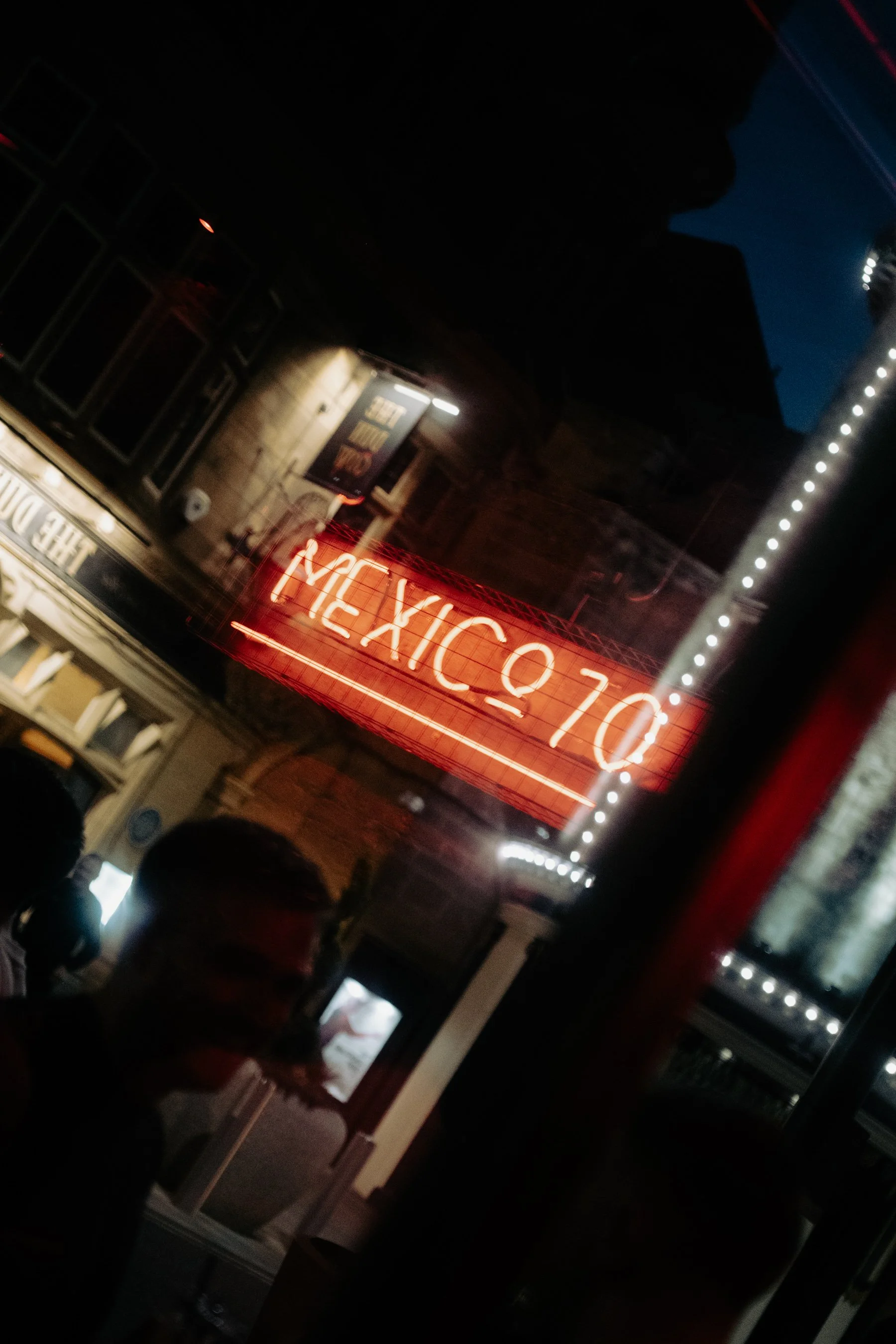Nighttime street scene with illuminated neon sign reading 'MEXICO' and people standing beneath it, buildings with lit windows, and string lights hanging overhead.
