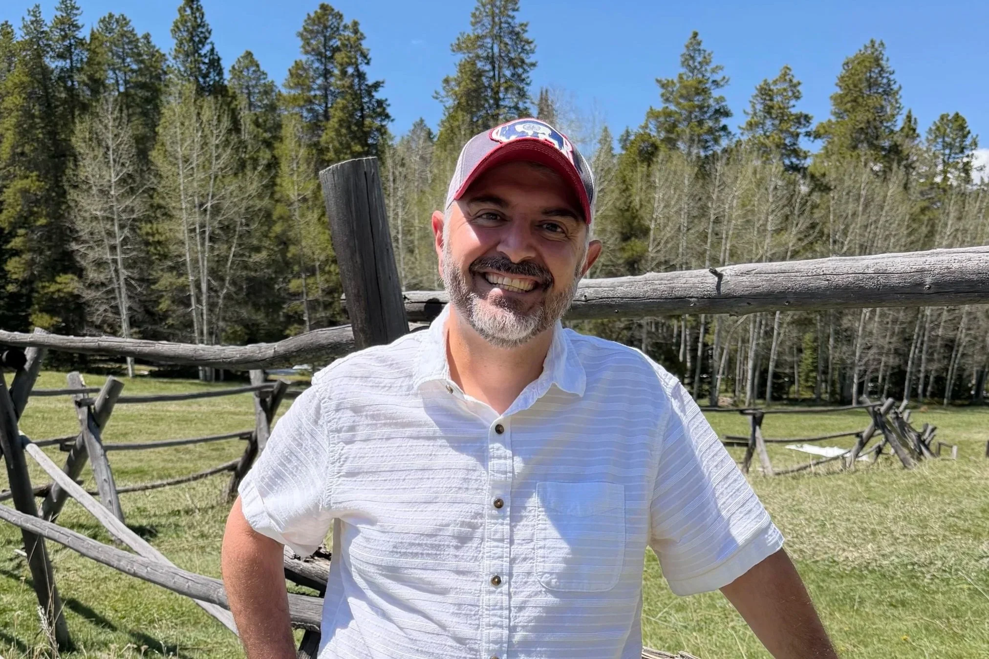 Ray Pacheco, Mayor of Casper and candidate for Natrona County Commissioner, standing in front of a wooden fence with Casper Mountain in the background. He is wearing a ball cap and tee shirt.