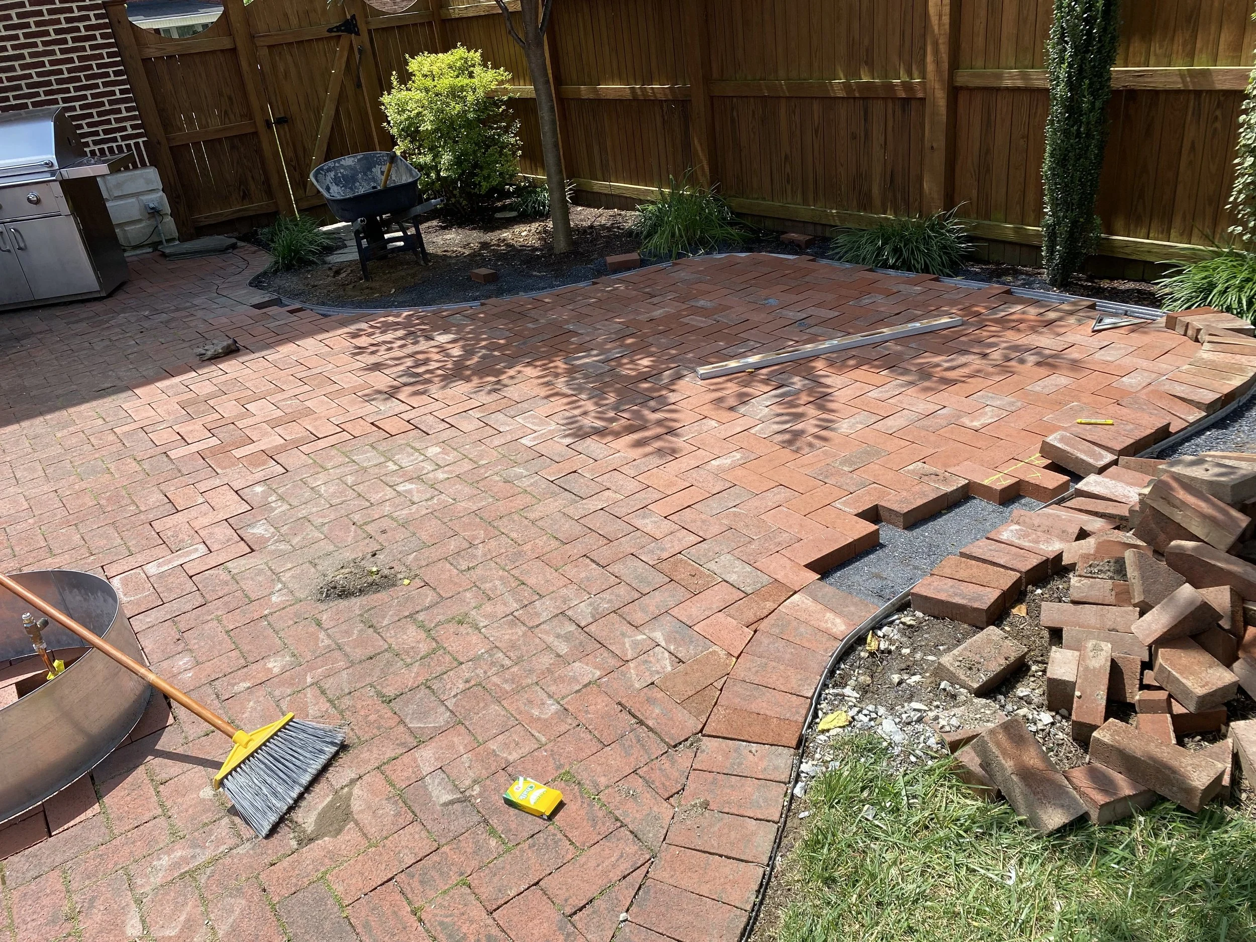 A backyard area with a newly laid brick patio, a wooden fence, and a small garden bed with trees and plants. Construction tools, including a broom and a level, are on the patio, and there are bricks and construction materials nearby.