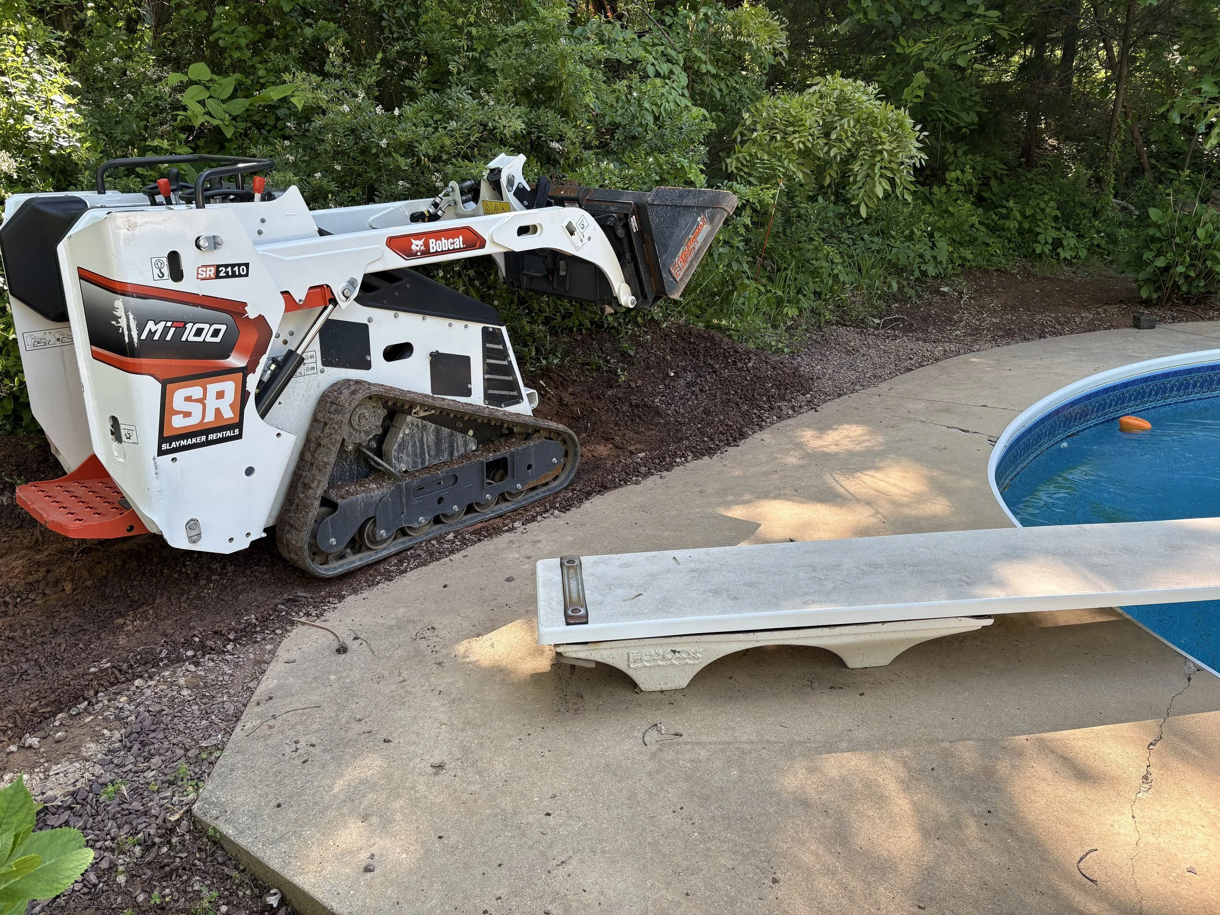 A Bobcat mini excavator working on landscaping next to a concrete patio, with plants and bushes in the background.