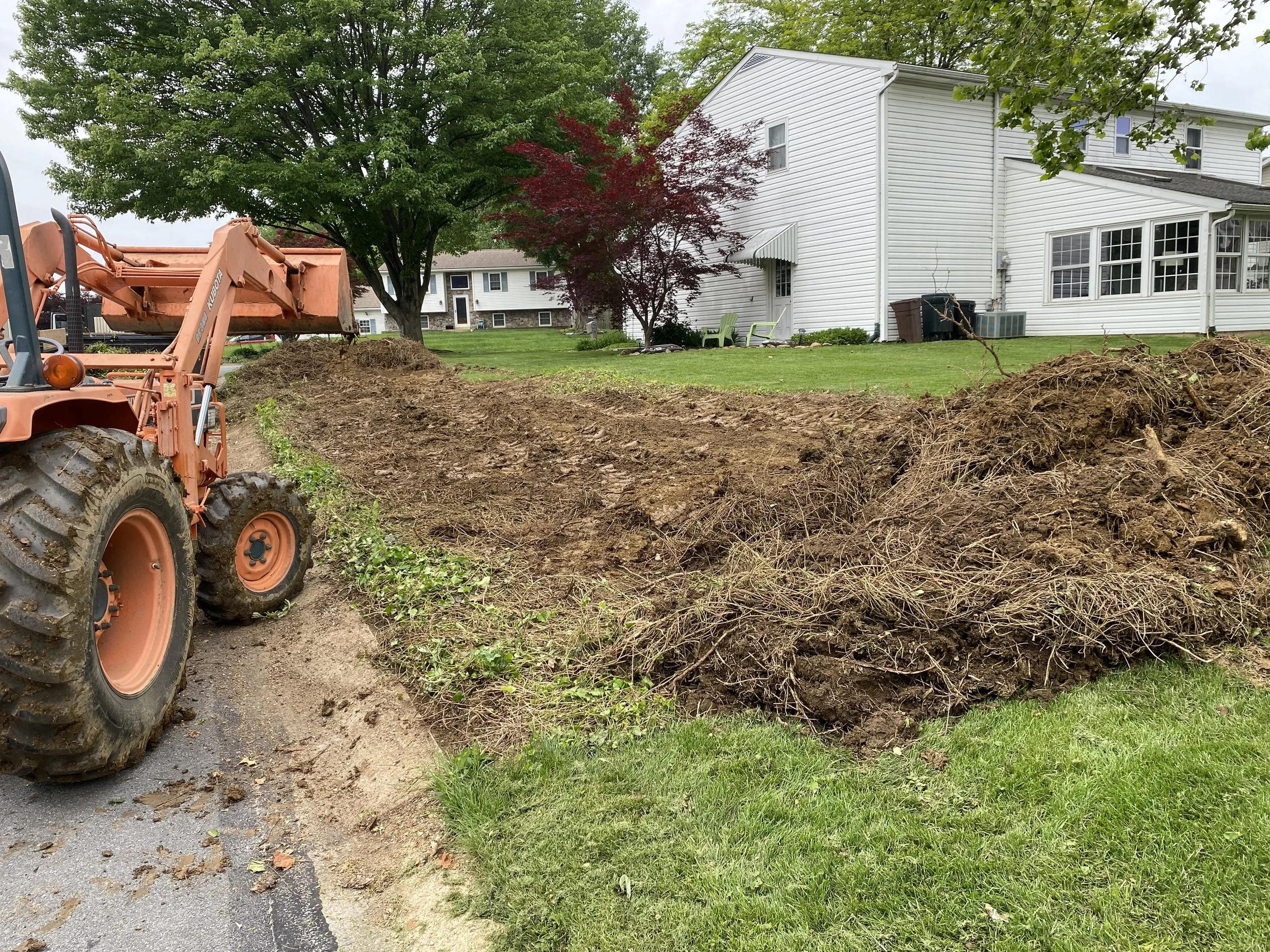 A construction vehicle is digging up soil near a grassy yard in front of a white house with large windows. Trees with green and red leaves are seen in the background.