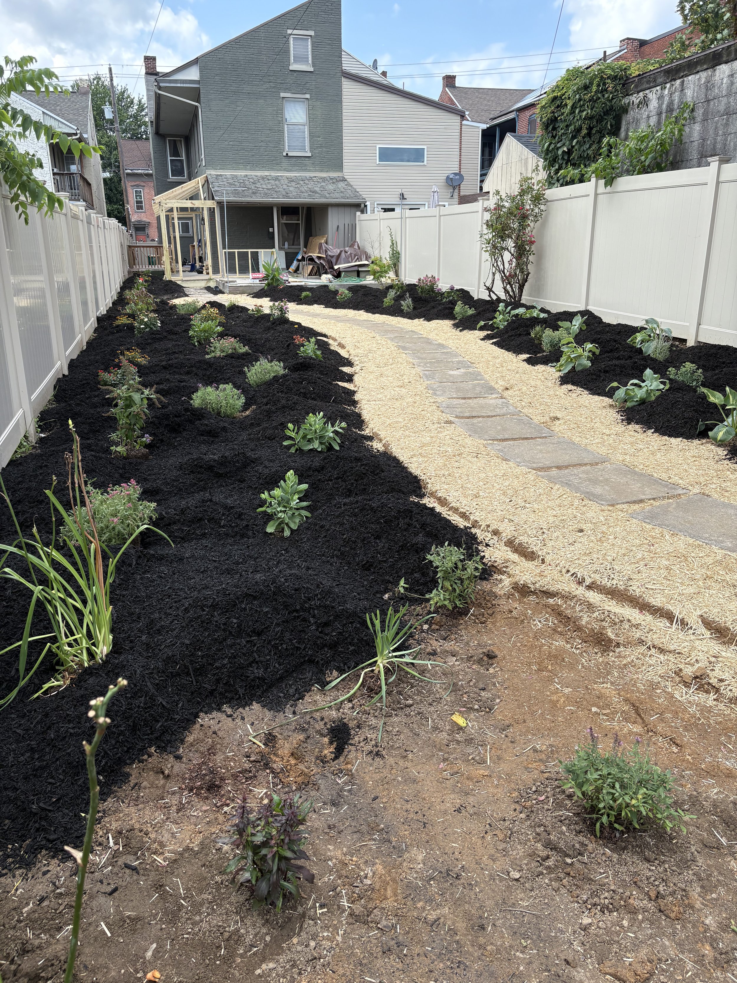 Backyard garden with newly planted flowers and shrubs, a curved gravel pathway, and a partially constructed wooden structure against a fence, with houses in the background.