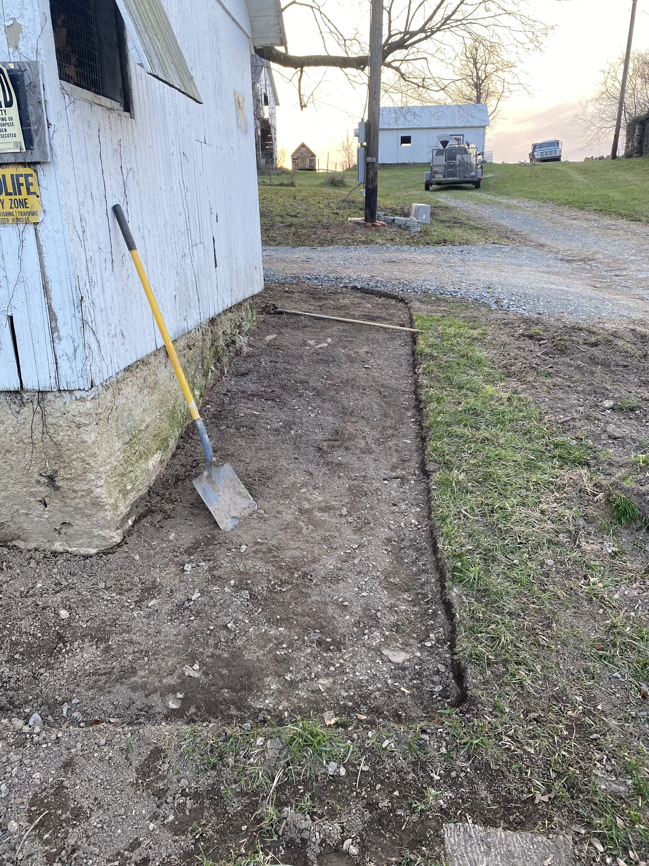 A partially dug trench alongside the side of a white painted building. There is a yellow-handled shovel with a silver blade leaning against the building. In the background, there is a gravel driveway, a grassy lawn, a white shed, and a utility traile