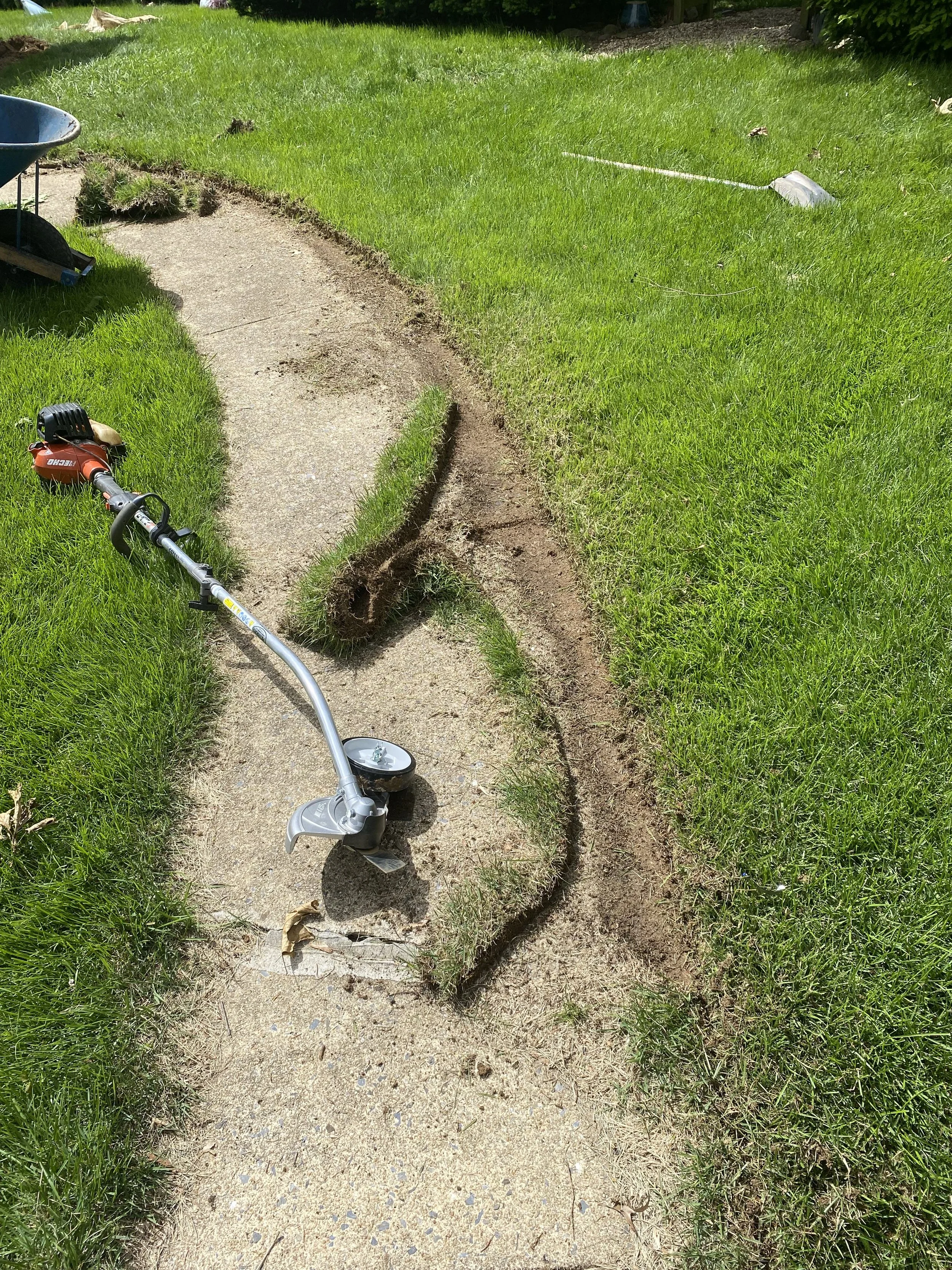 A garden pathway is being replaced, with a section of concrete removed and a string marking the edge of the new grass area. A string line tool and a grass trimmer are visible on the ground.