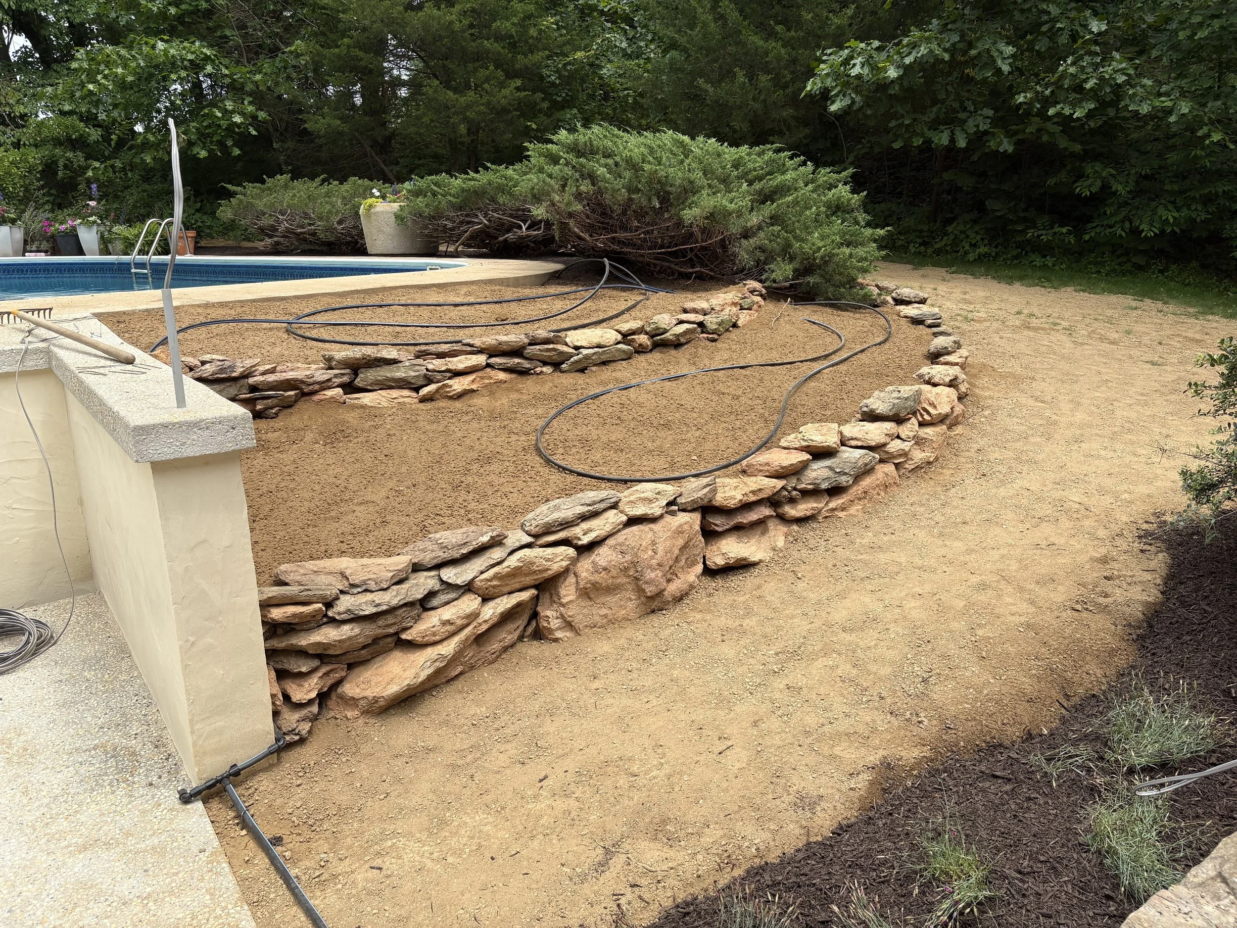 Construction site with a stone retaining wall, a partially completed concrete structure, and a garden with plants in pots. Soil and landscaping work in progress with garden hoses laid out.