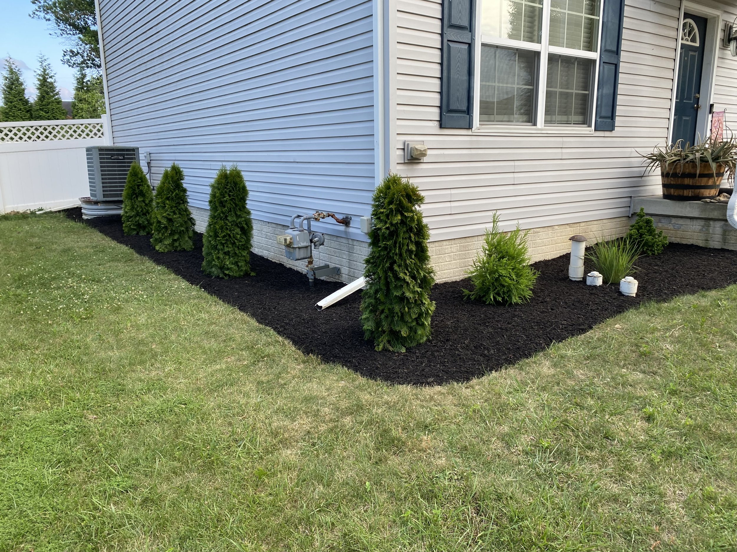 A house side with white vinyl siding, blue shutters, a window, a white vinyl fence, small trees, an air conditioning unit, and a garden with plants and mulch.