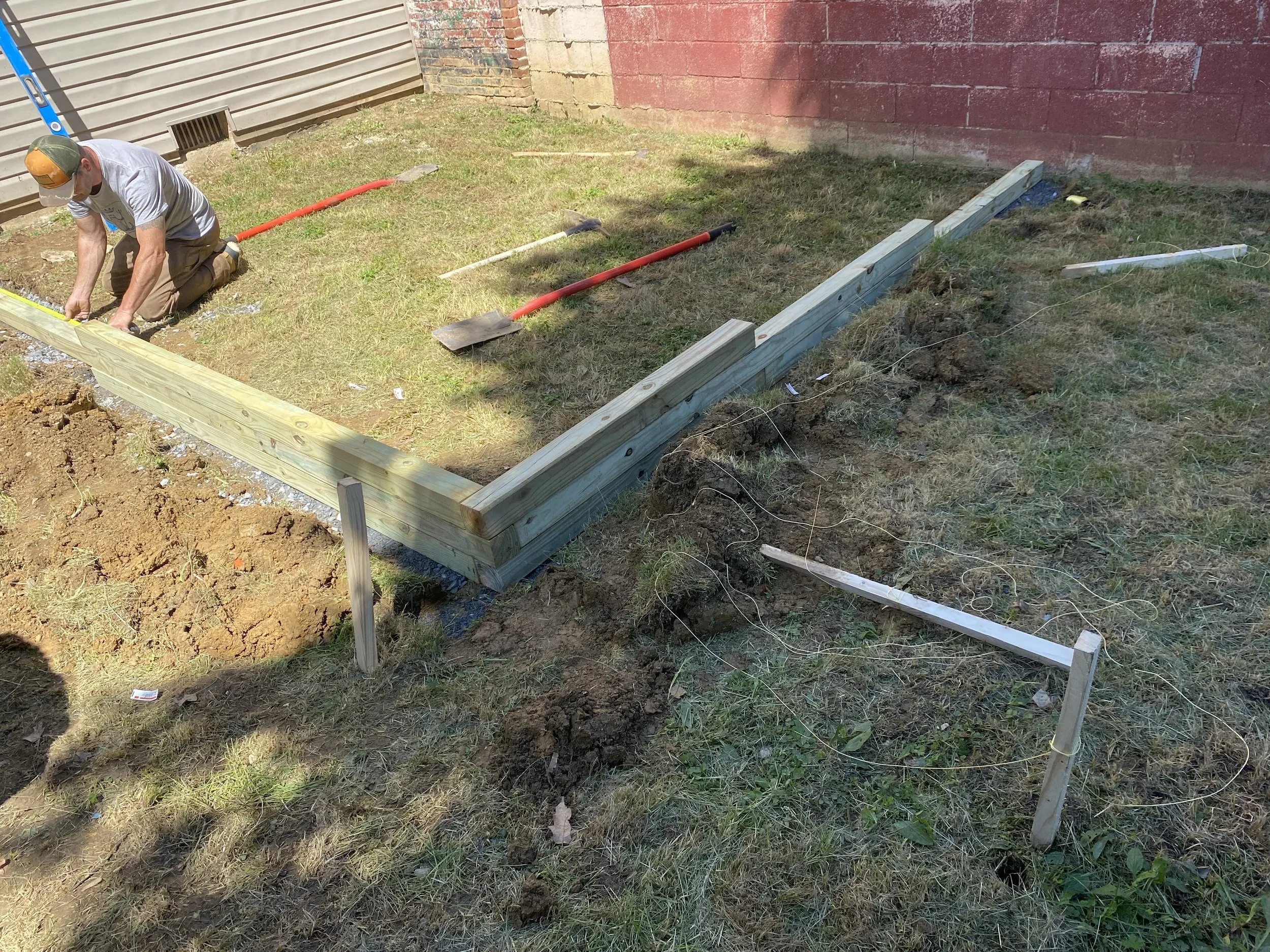 A man kneeling on the grass working on a construction project, installing a wooden frame for a small wall or foundation, with various tools and materials scattered around.