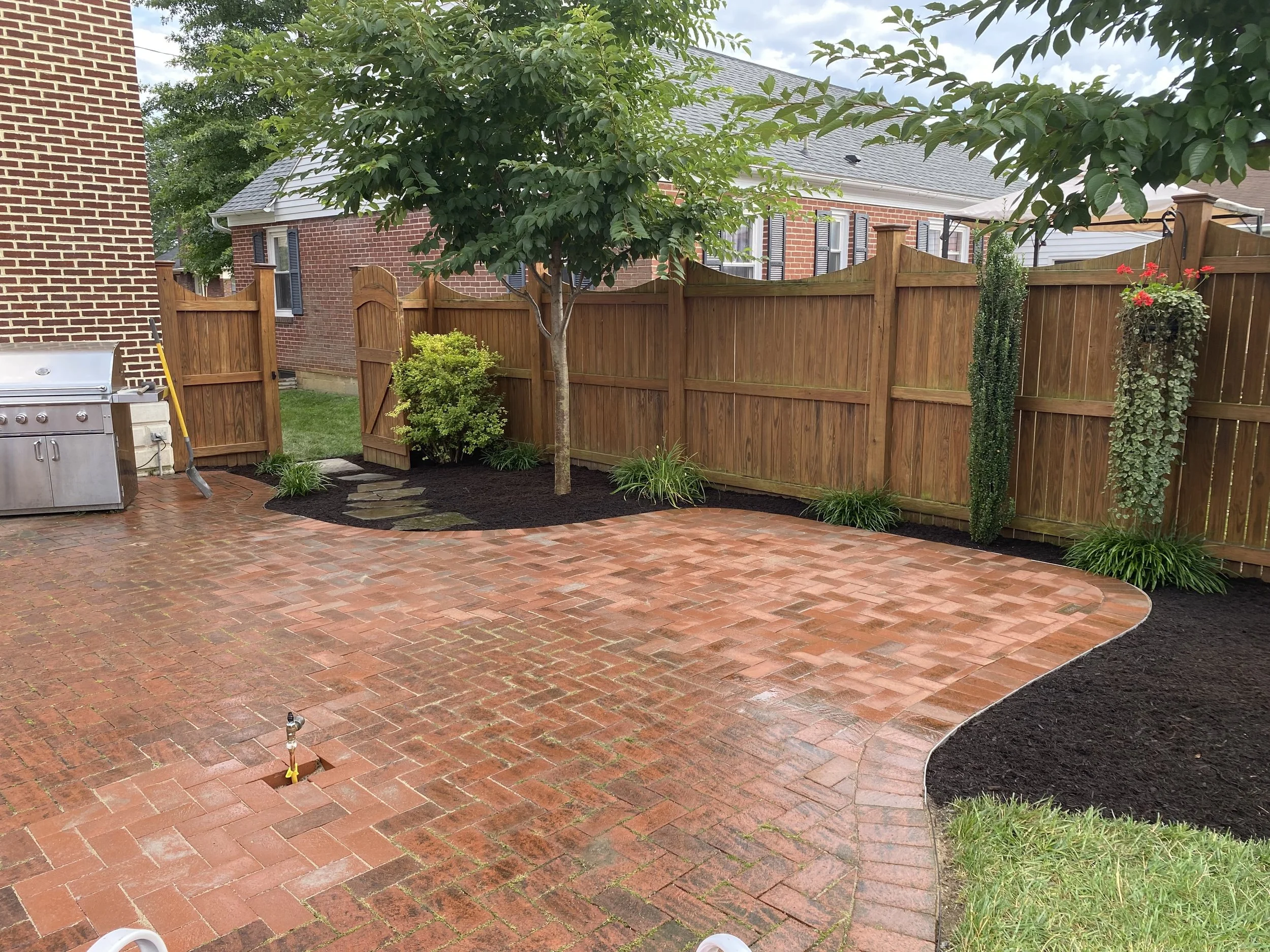 Backyard with brick patio, small trees, flowered hanging baskets, and a tall wooden fence.