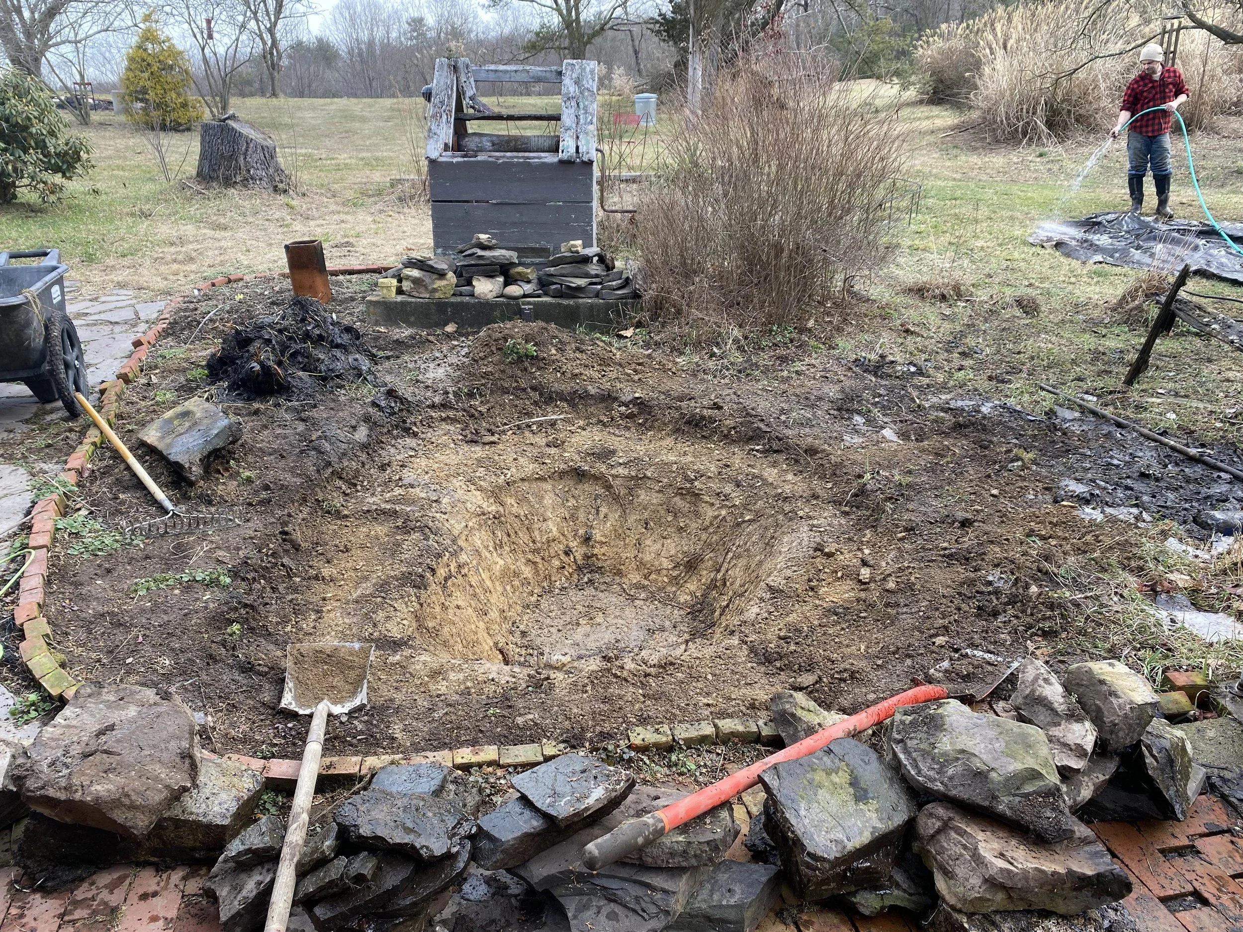 A person in a red and black plaid shirt and boots is watering a garden bed with a hose. There is a sunken area in the ground with dirt, rocks, and some garden tools, and a stone border around part of the garden. The setting appears to be a yard or ou