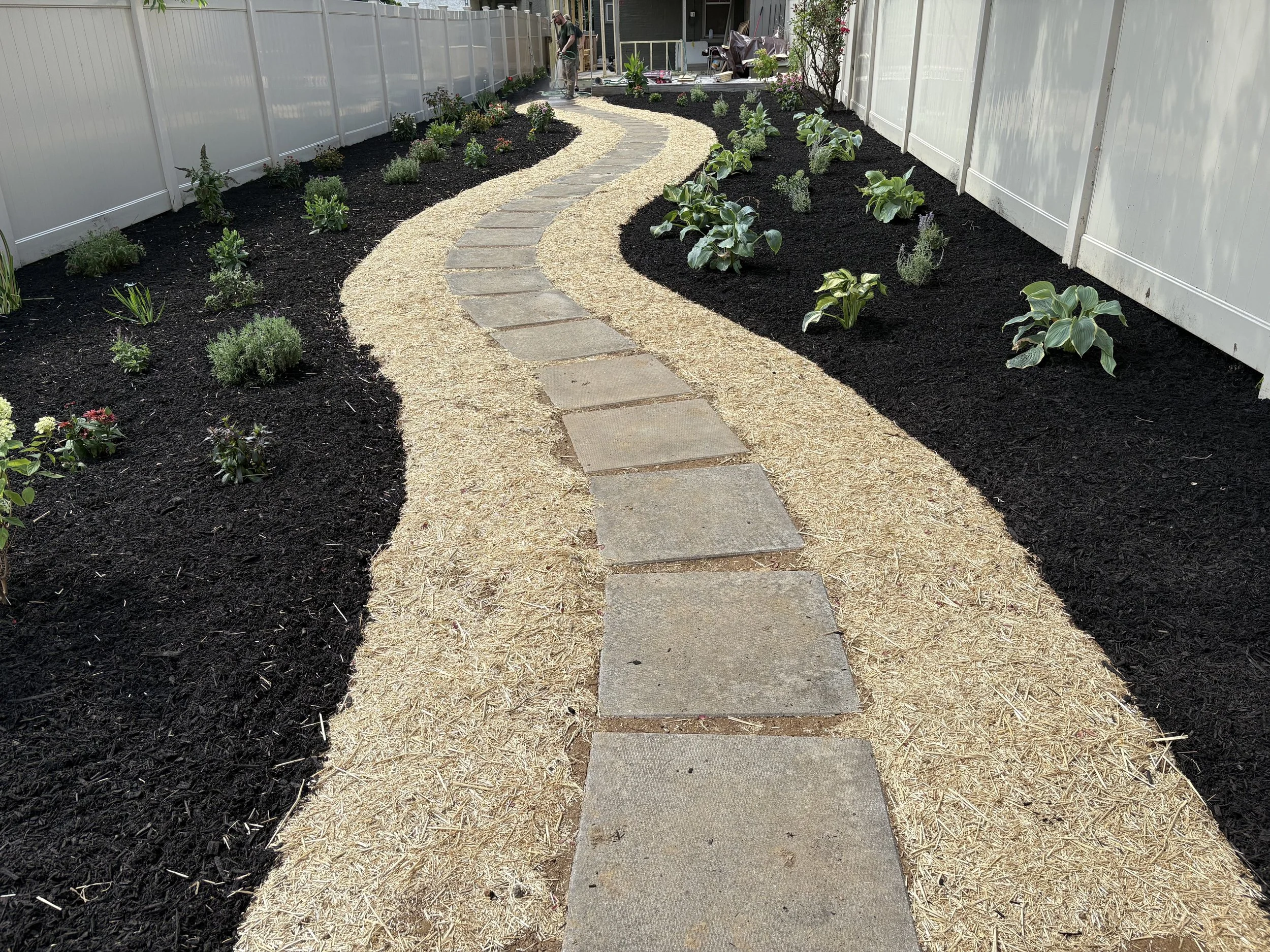 A backyard garden with a winding stone pathway, black mulch on the sides, and various green plants. A person is working near a house in the background.