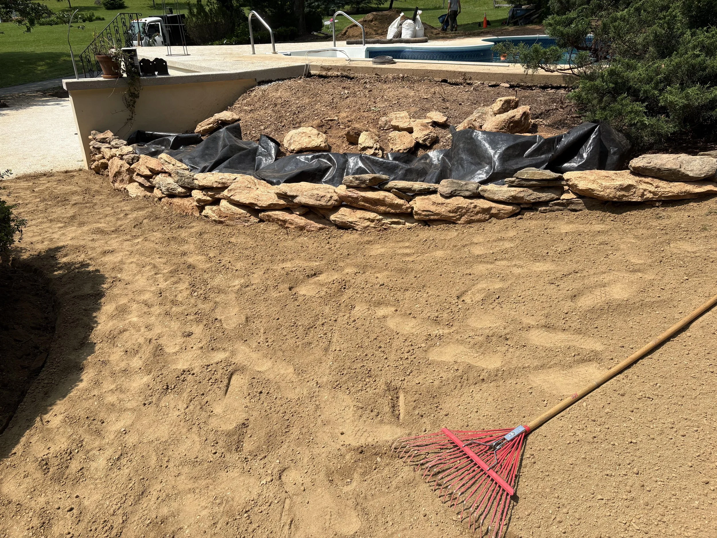 A backyard pool area with a section of a garden bed under construction. The garden bed has rocks along the edge, black plastic liner, and exposed soil. A red rake and a landscape tool are lying on the soil. In the background, there is a pool, a ladde