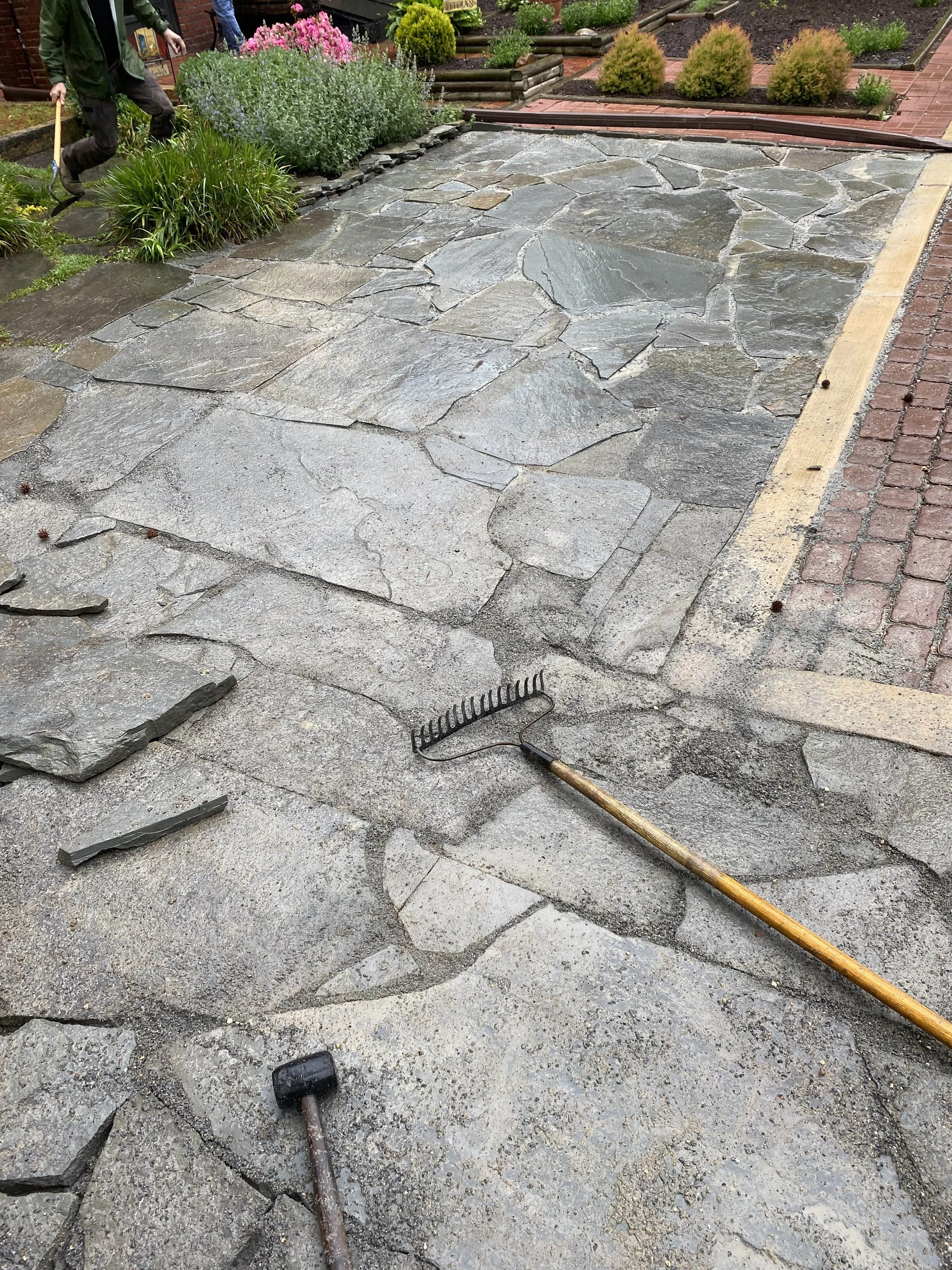 The image shows a stone-paved outdoor area with some gardening tools, including a rake and a hammer, placed on the ground. There are some plants and a brick walkway adjacent to the stone pavement. The area appears to be under maintenance or renovatio