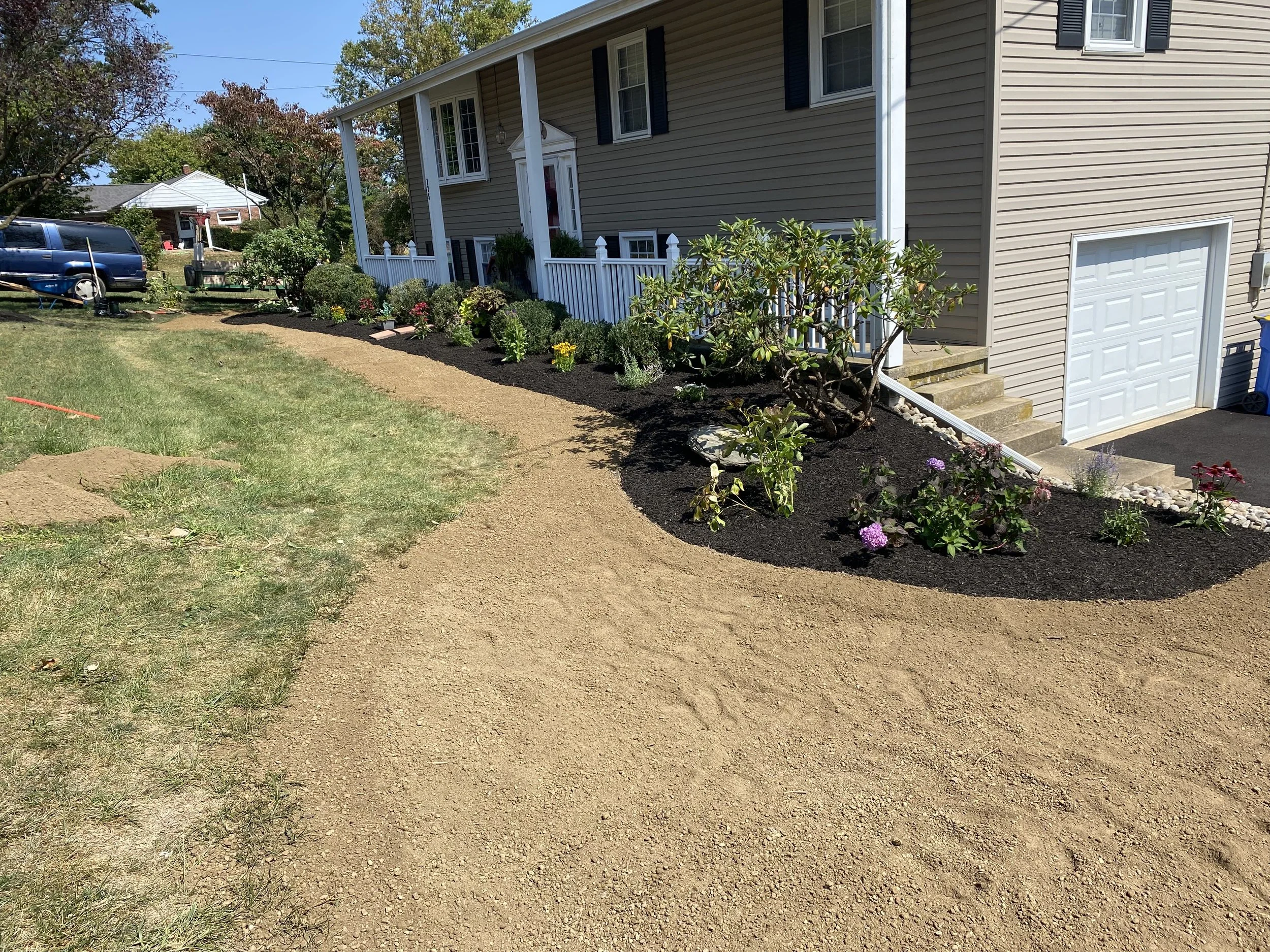 A residential backyard with a beige house, white picket fence, garden bed with blooming flowers, and a gravel pathway.