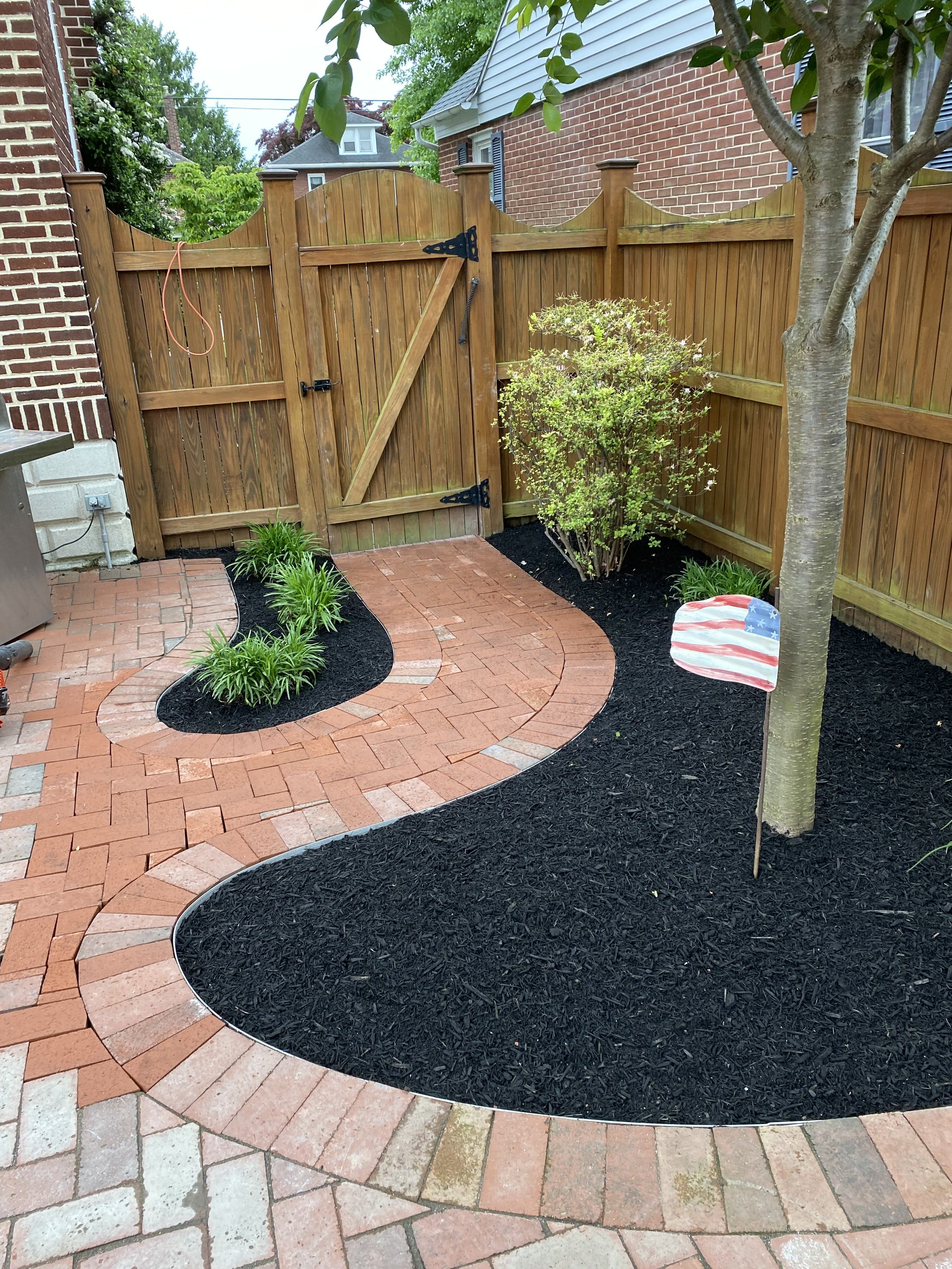 A backyard with a brick pathway, garden bed with green plants, a wooden fence, and a small American flag decoration. There are trees and houses visible in the background.