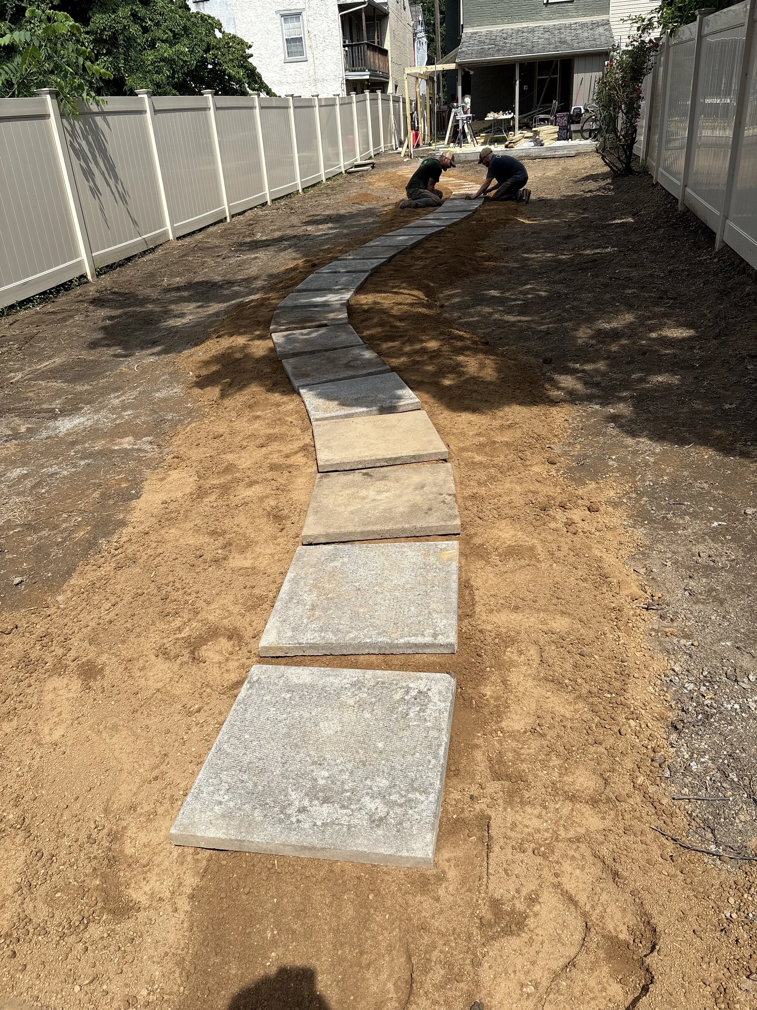 Two men are kneeling and working on a curved pathway of concrete stepping stones in a backyard, with a white fence on each side.