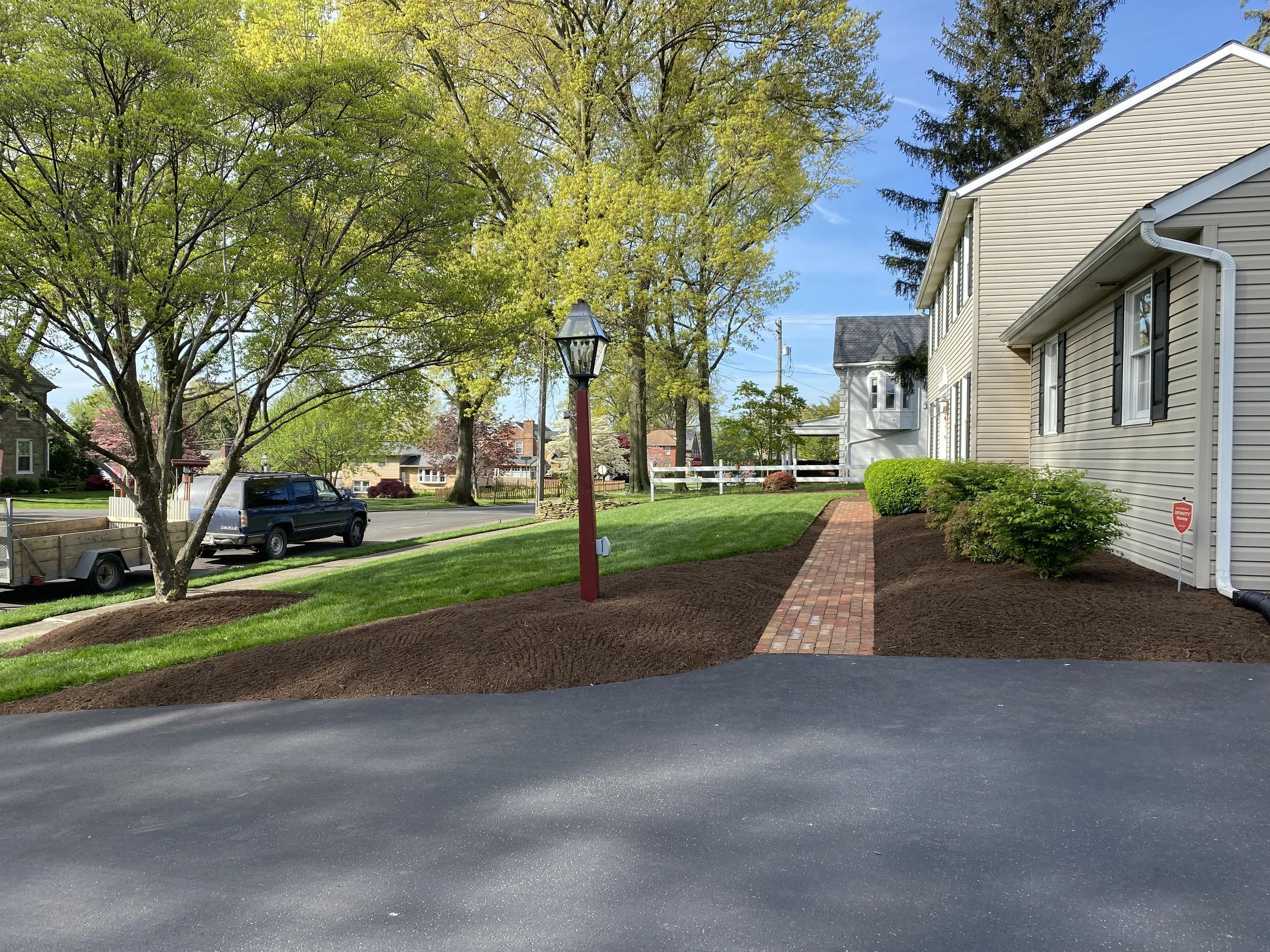 A suburban street with green trees, a parked vehicle, and residential houses under a blue sky. A brick sidewalk lines the property next to a manicured lawn and shrubs.