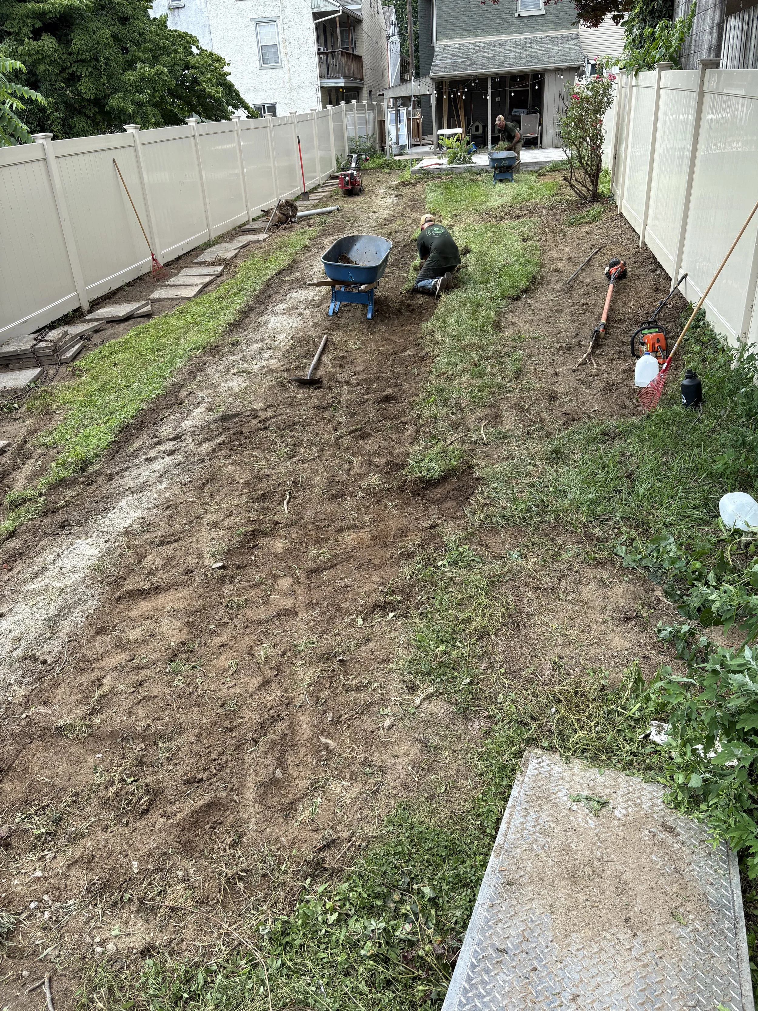 Home improvement workers landscaping and preparing soil in a backyard, with tools and wheelbarrows scattered around.