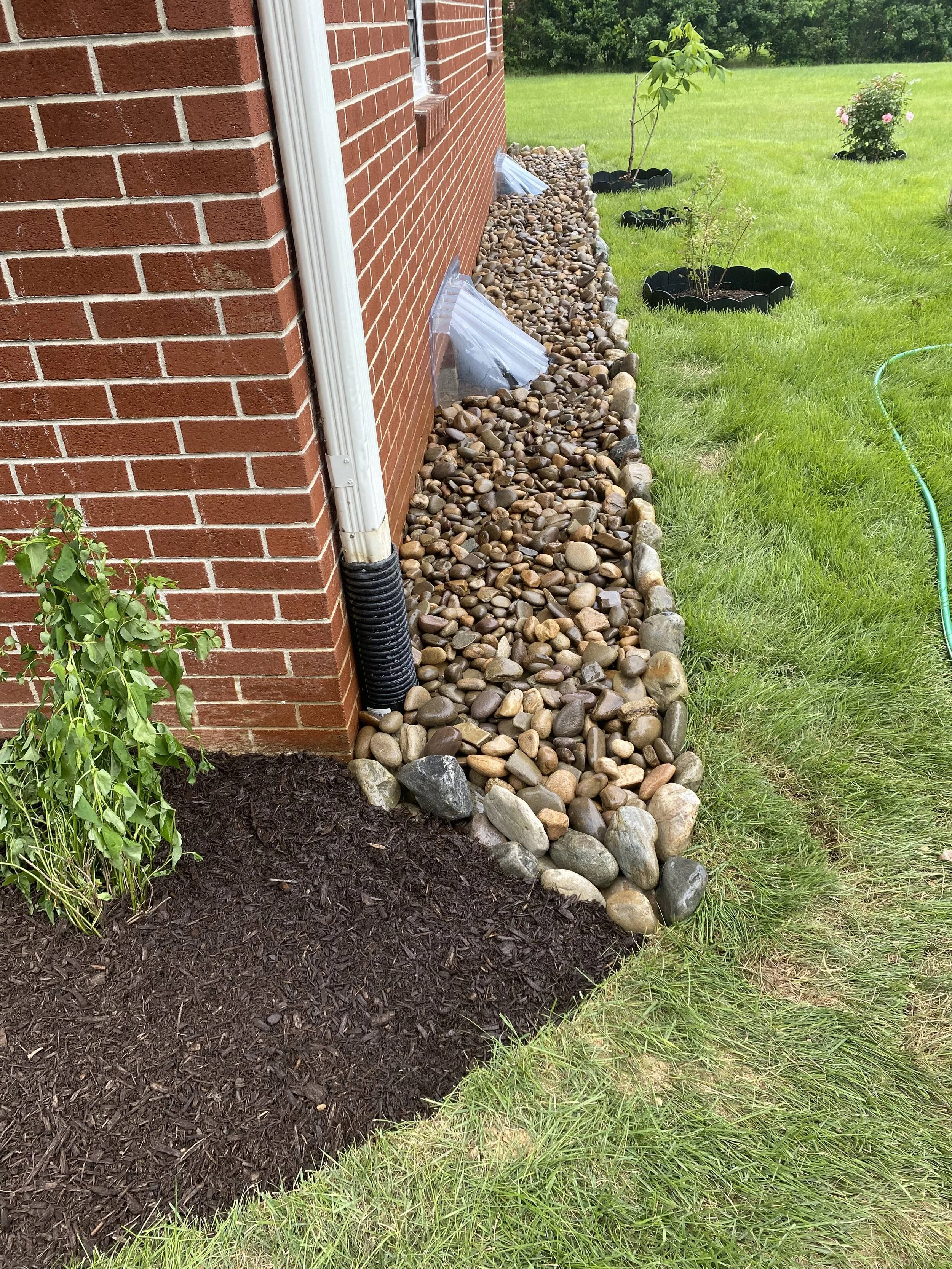 The image shows the side of a brick house with a layer of small rocks and stones along the foundation, next to a green lawn with flower beds and young plants, and a garden hose on the grass.