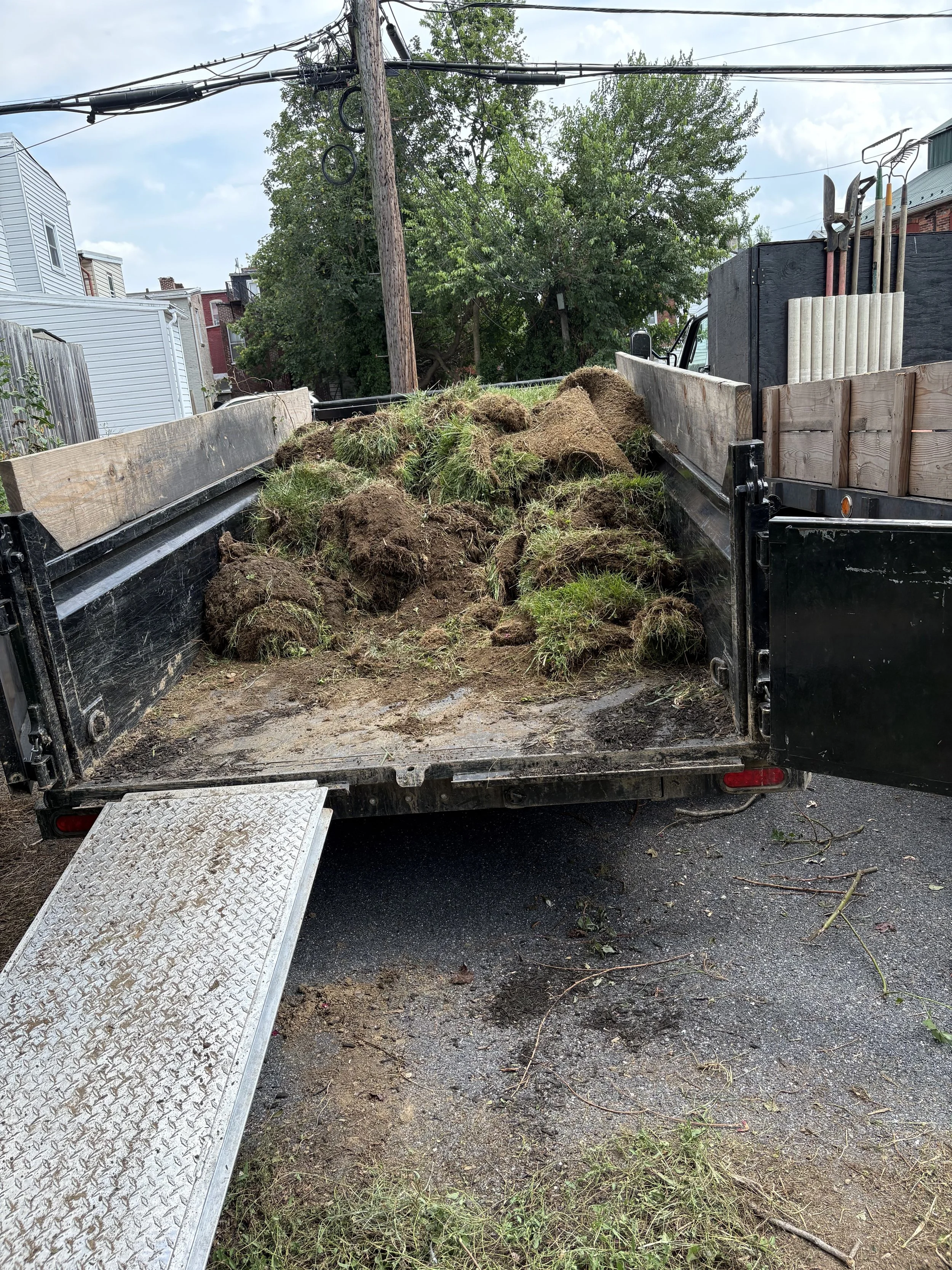 A flatbed truck filled with dirt, grass, and plant debris parked on a street near a residential area with houses, trees, and utility poles.