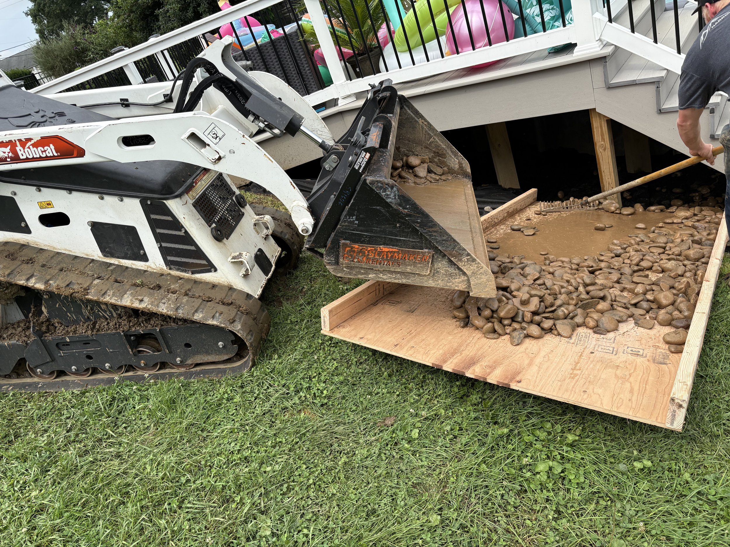 A person operating a small Bobcat skid-steer loader, moving large, smooth rocks into a wooden frame resembling a pathway or landscaping feature, next to a house with a deck and pool floaties visible in the background.