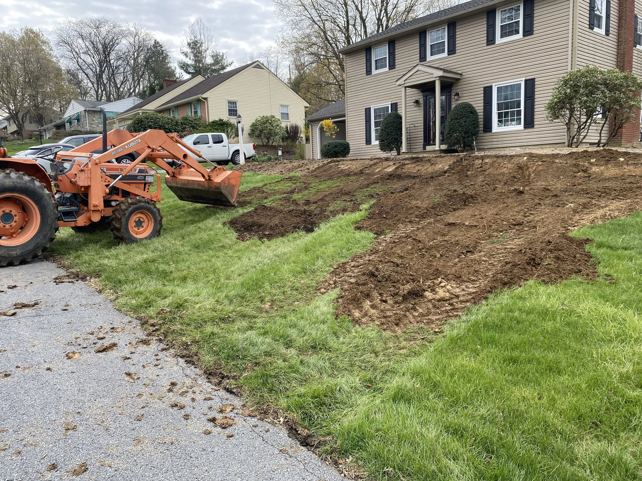 A backyard with a house, trees, and a tractor digging up the yard, creating a trench or garden bed.
