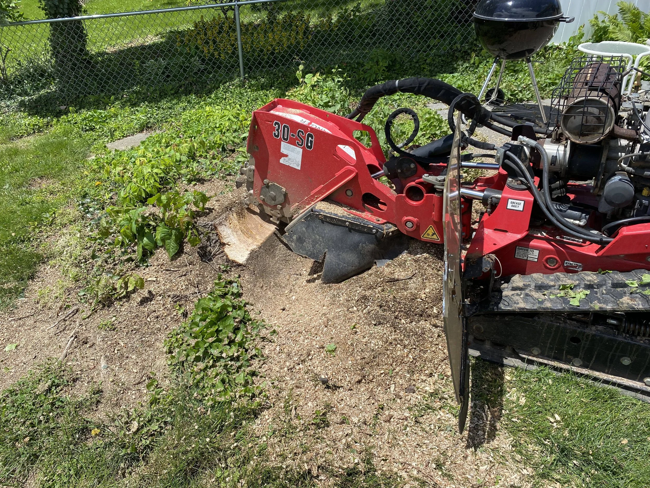 A red mini excavator digging in a backyard with a black grill and white chair in the background, surrounded by green grass and a chain-link fence.