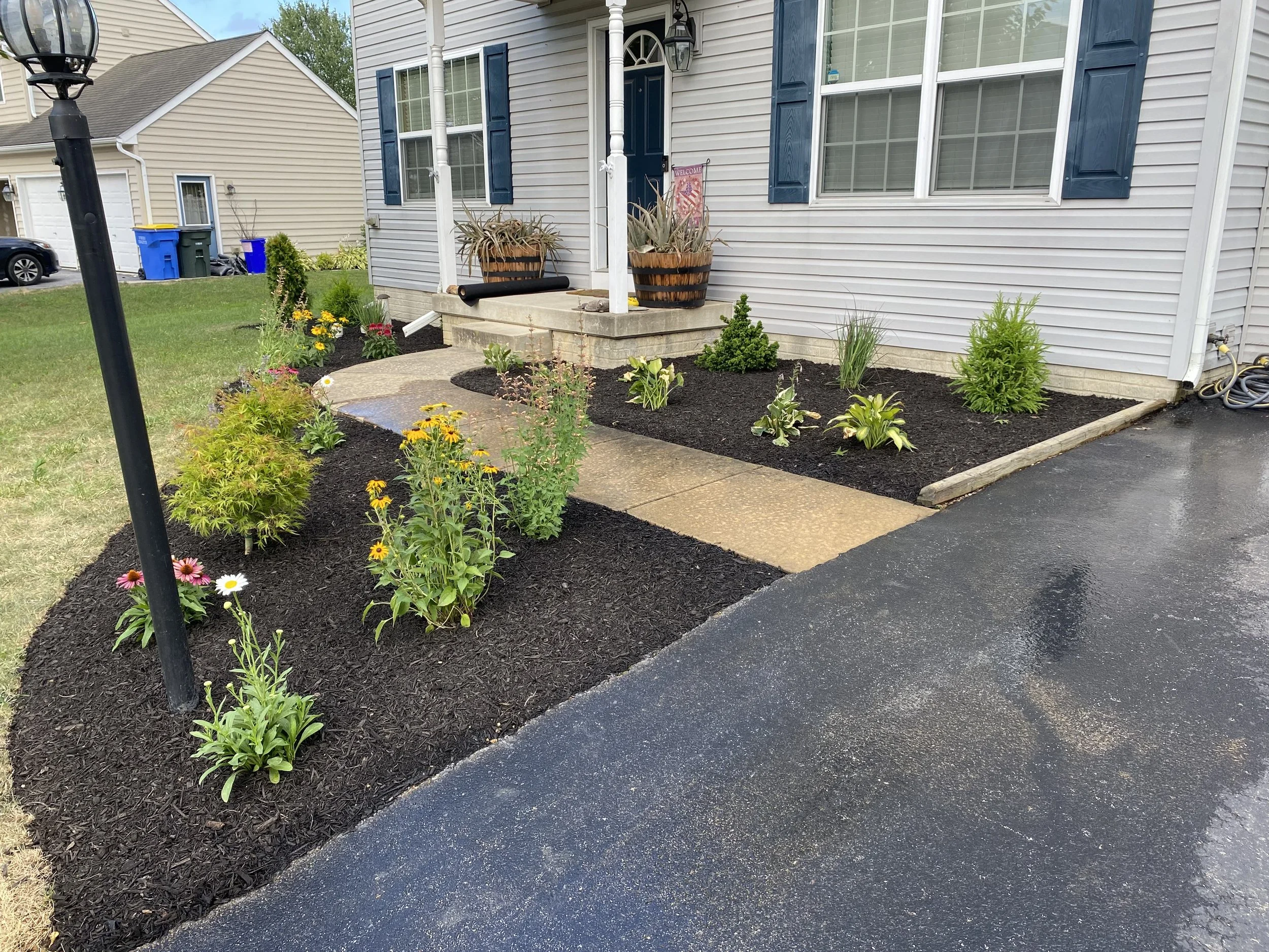 A house with light gray siding and blue shutters has a small front garden with flowers and plants, a concrete walkway, and a blacktop driveway.