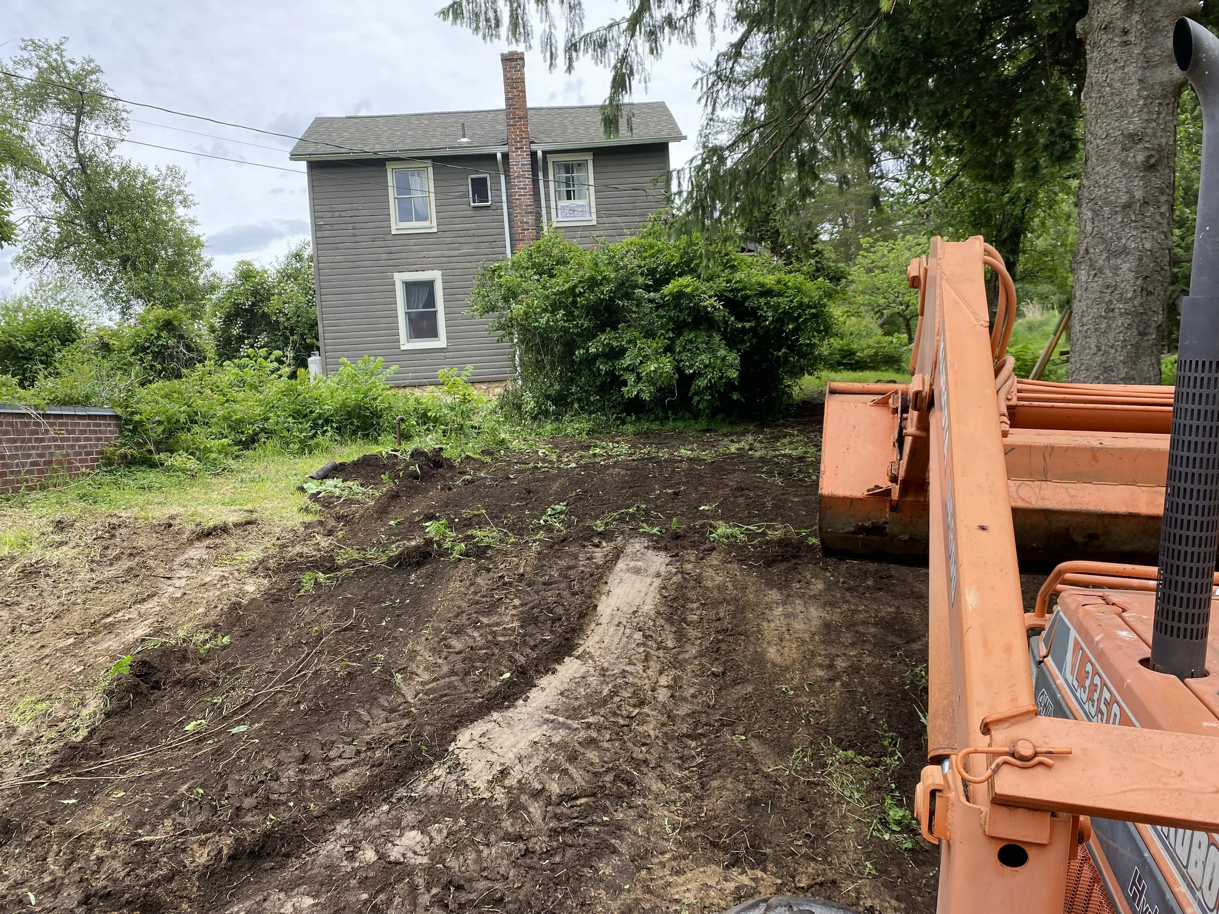 A partially excavated backyard with a gray house in the background, surrounded by green trees and bushes, and construction equipment in the foreground.
